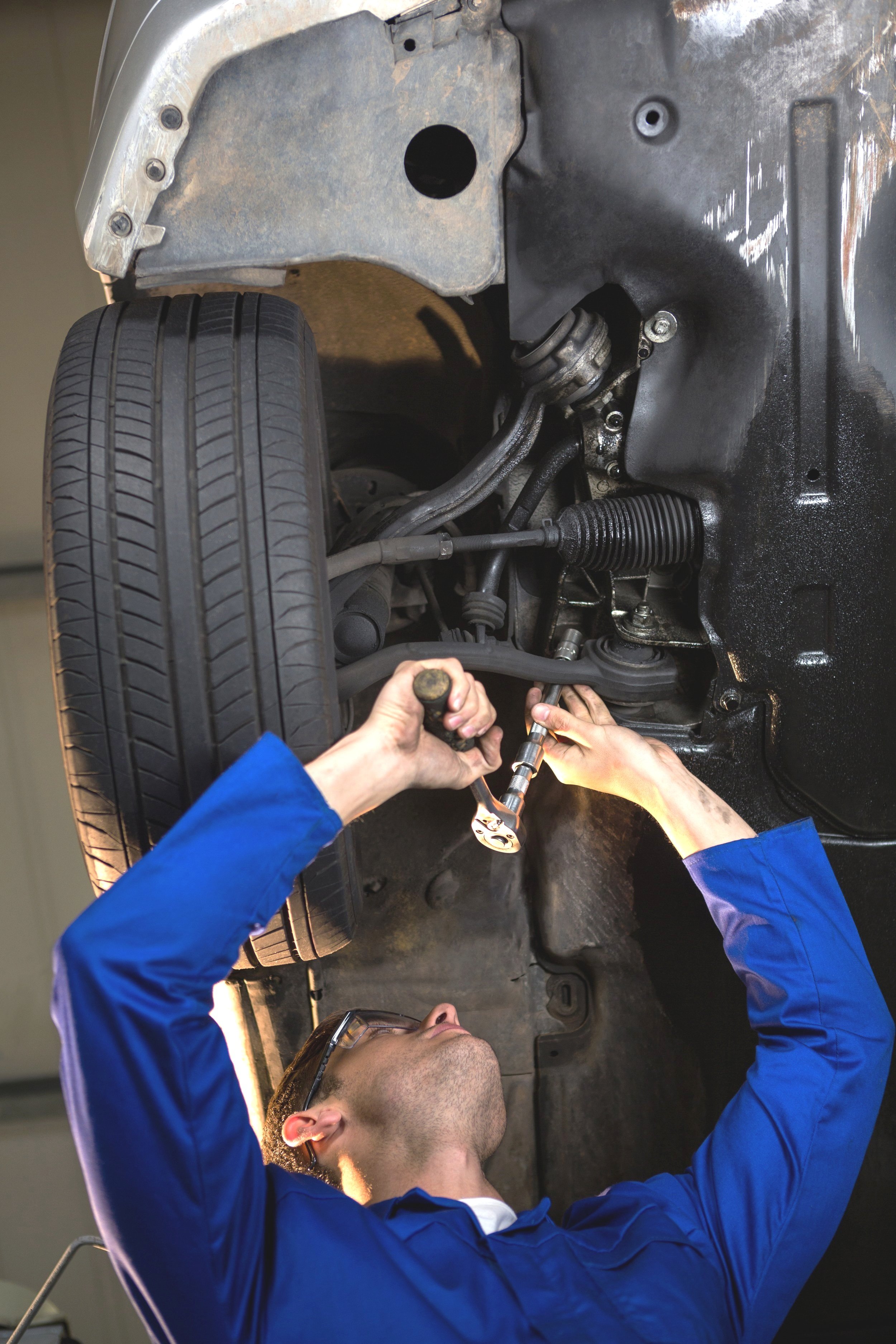 A mechanic working underneath a vehicle, using tools to repair or maintain parts of the car's suspension or steering system.
