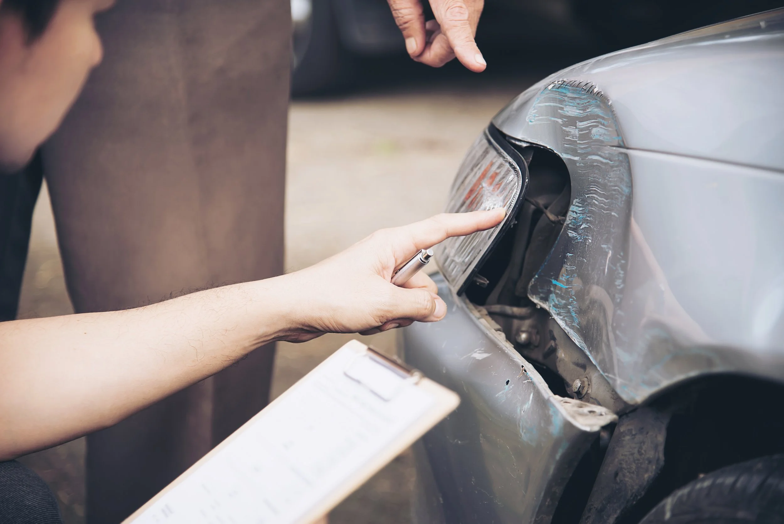 Person inspecting front bumper and headlight of a damaged silver car, holding a pen and clipboard near the scene of an accident.