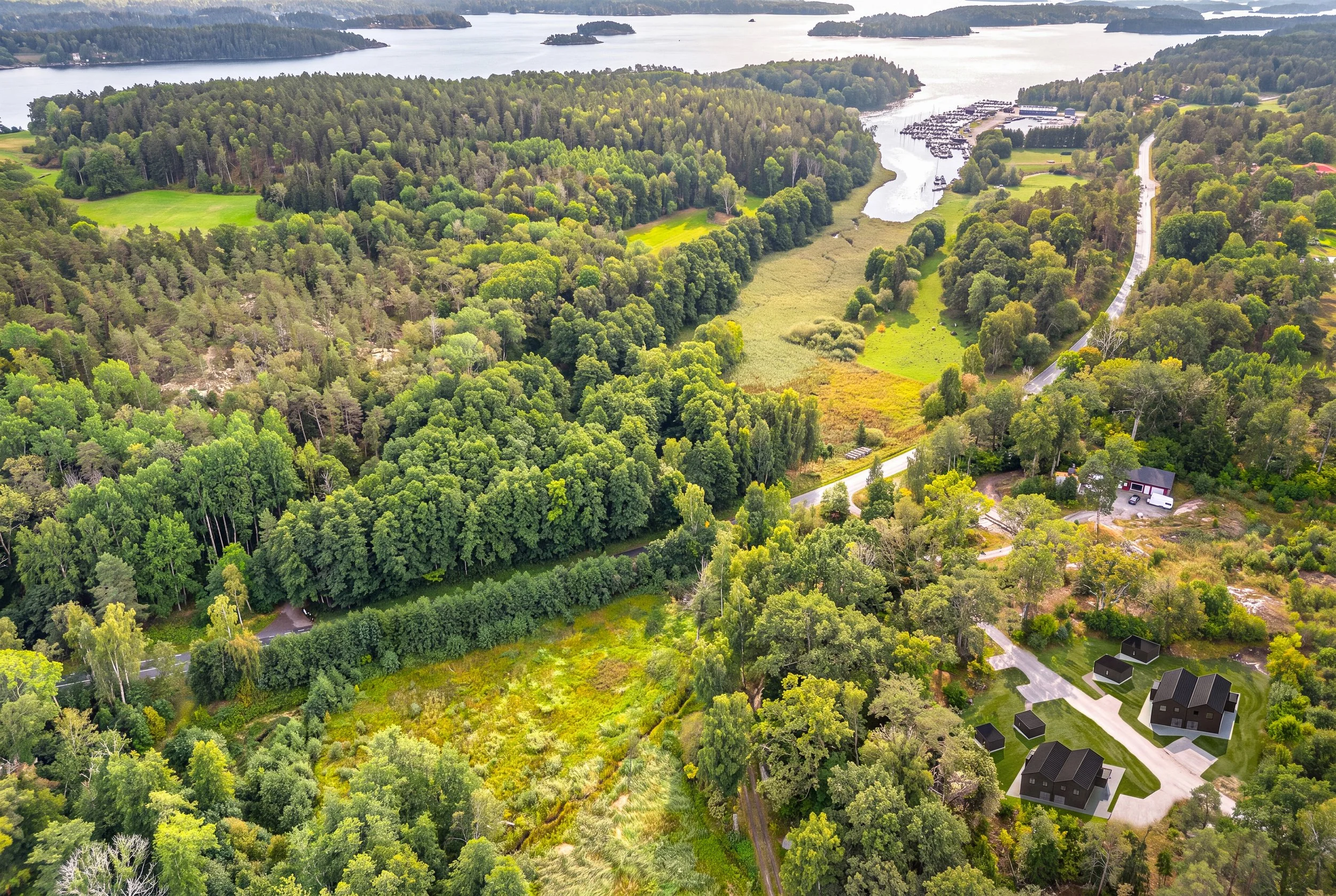 Flygbild av ett grönt skogsområde med några hus och en närliggande sjö eller vatten.