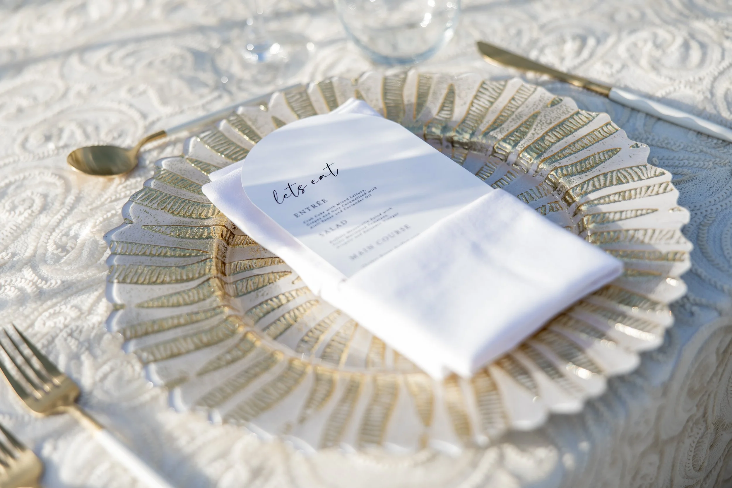 Mesa de evento elegante con un plato decorativo dorado y blanco, un menú en papel y cubiertos de oro, en una mesa con mantel de encaje.