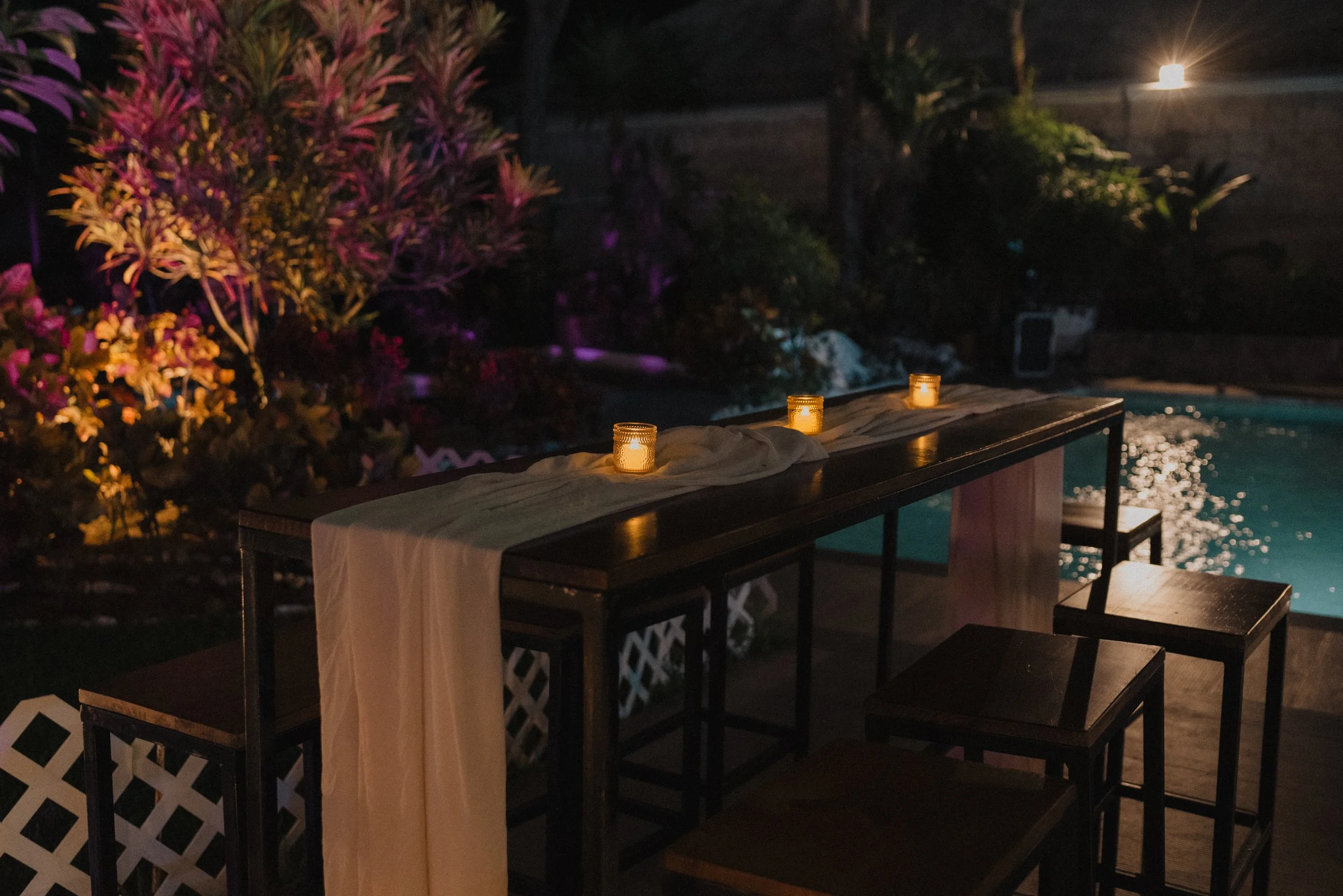 Mesa decorada con velas junto a una piscina en una noche oscura, con plantas y luces en el fondo.