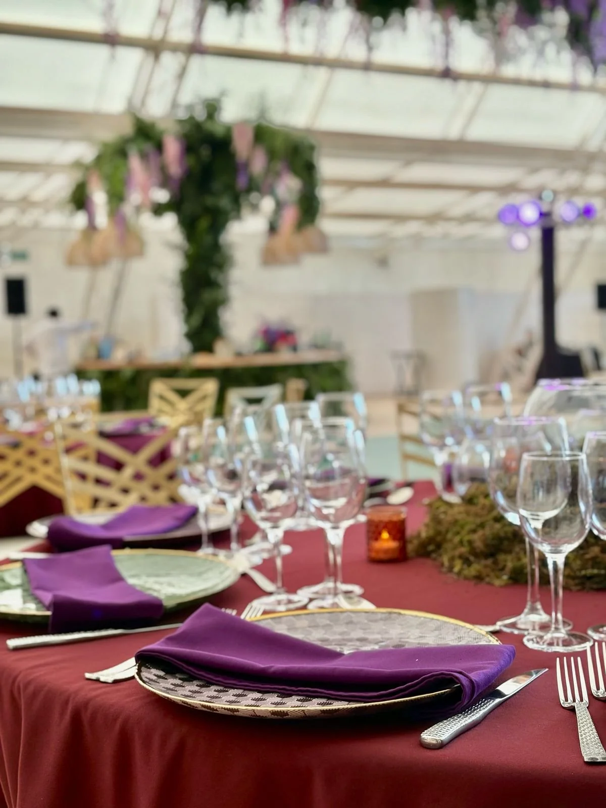 Mesa decorada para evento con manteles rojos, cubiertos plateados, copas, platos con servilletas moradas, y fondo con decoración floral y luces