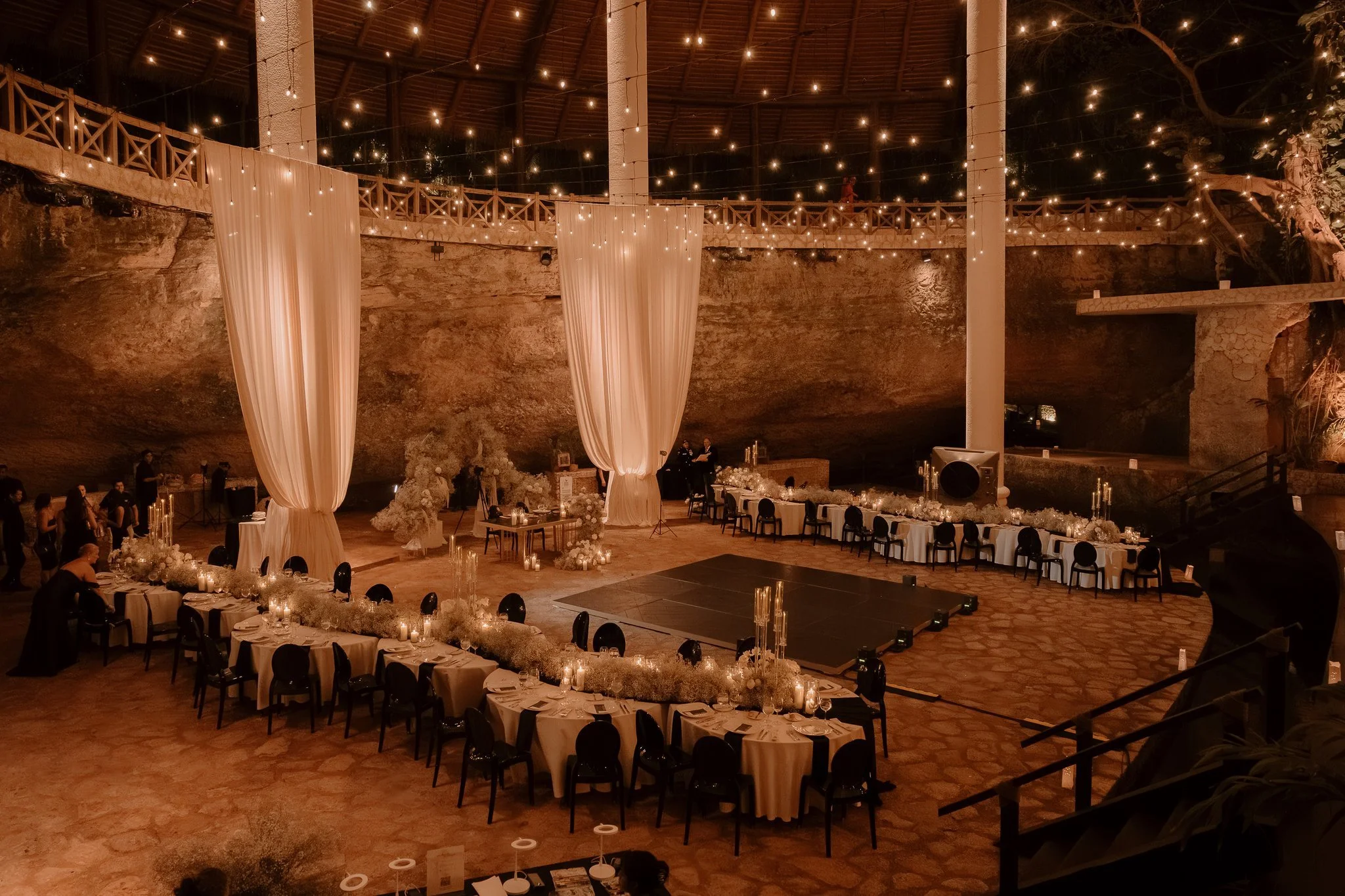 Decoración elegante para evento en un salón con techo alto, cortinas blancas, luces colgantes y mesas con velas y arreglos florales.