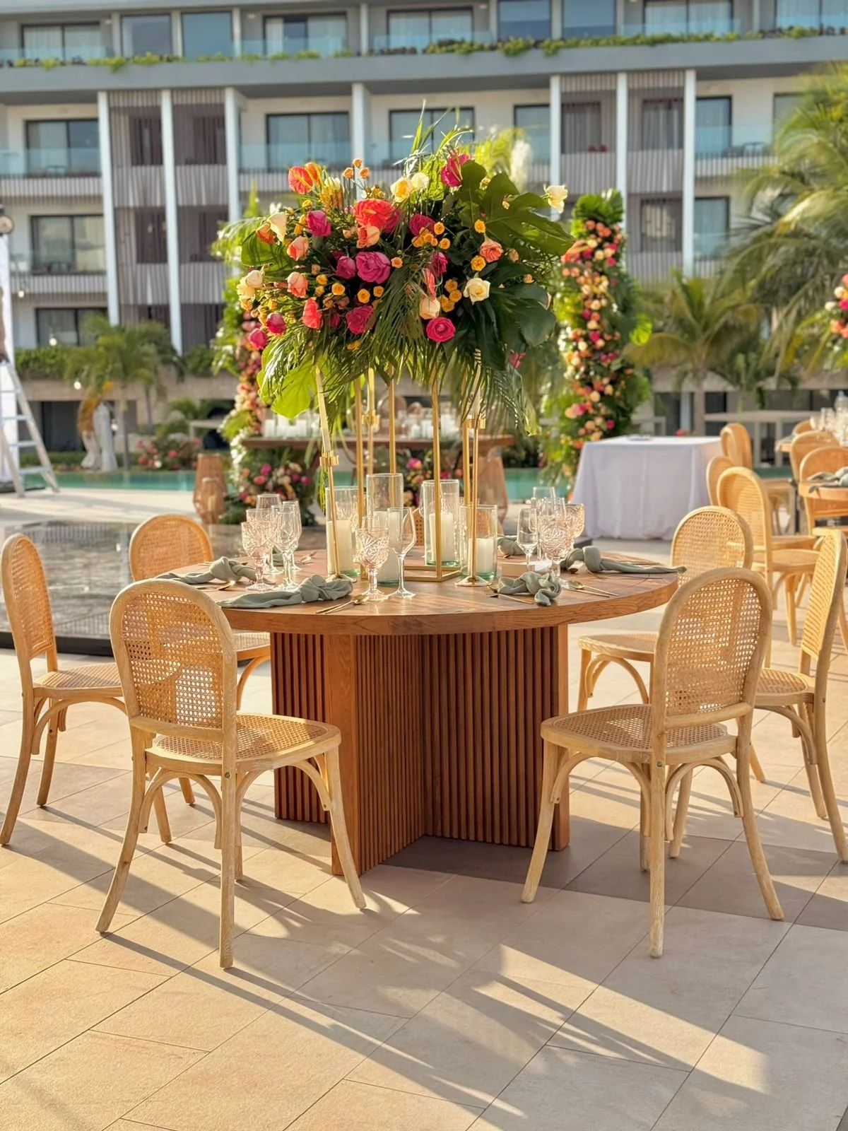 Mesa decorada con centro de flores coloridas y copas de cristal, en un evento junto a una alberca y edificio moderno.