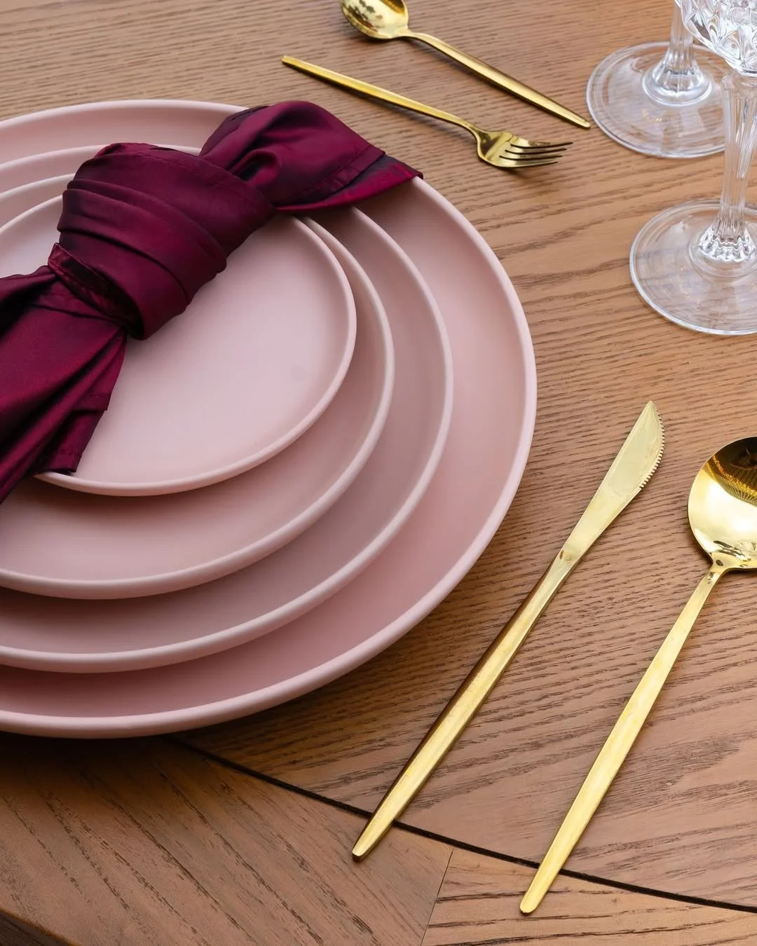 Configuración de mesa con platos rosas, servilleta morada, cubiertos dorados y copas de cristal sobre mesa de madera.