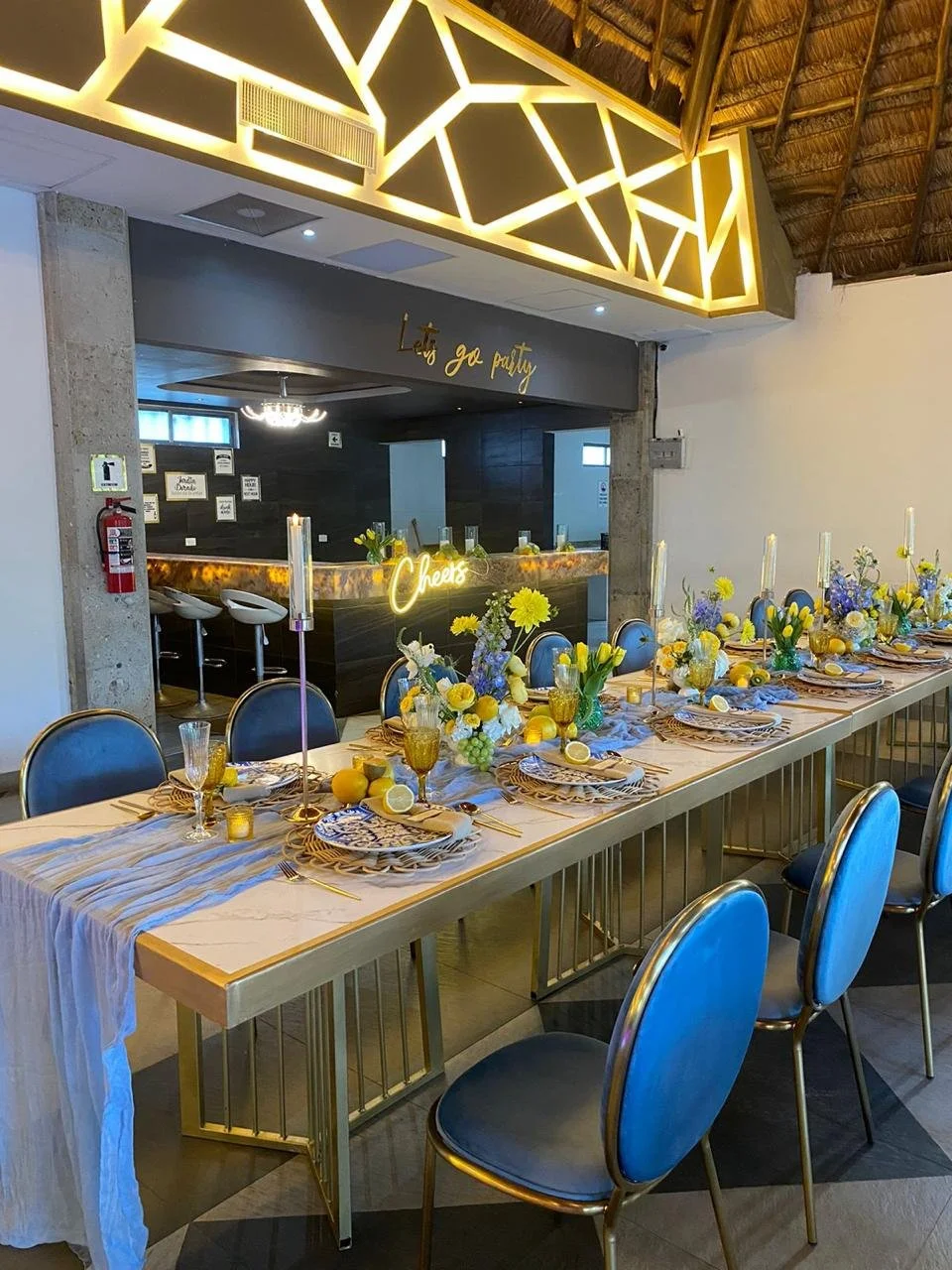 Mesa decorada para una fiesta con flores amarillas y azules, manteles y platos con temática cítrica, decoraciones de velas altas y un cartel que dice "Let's go party" en un restaurante con diseño moderno y techo de paja.