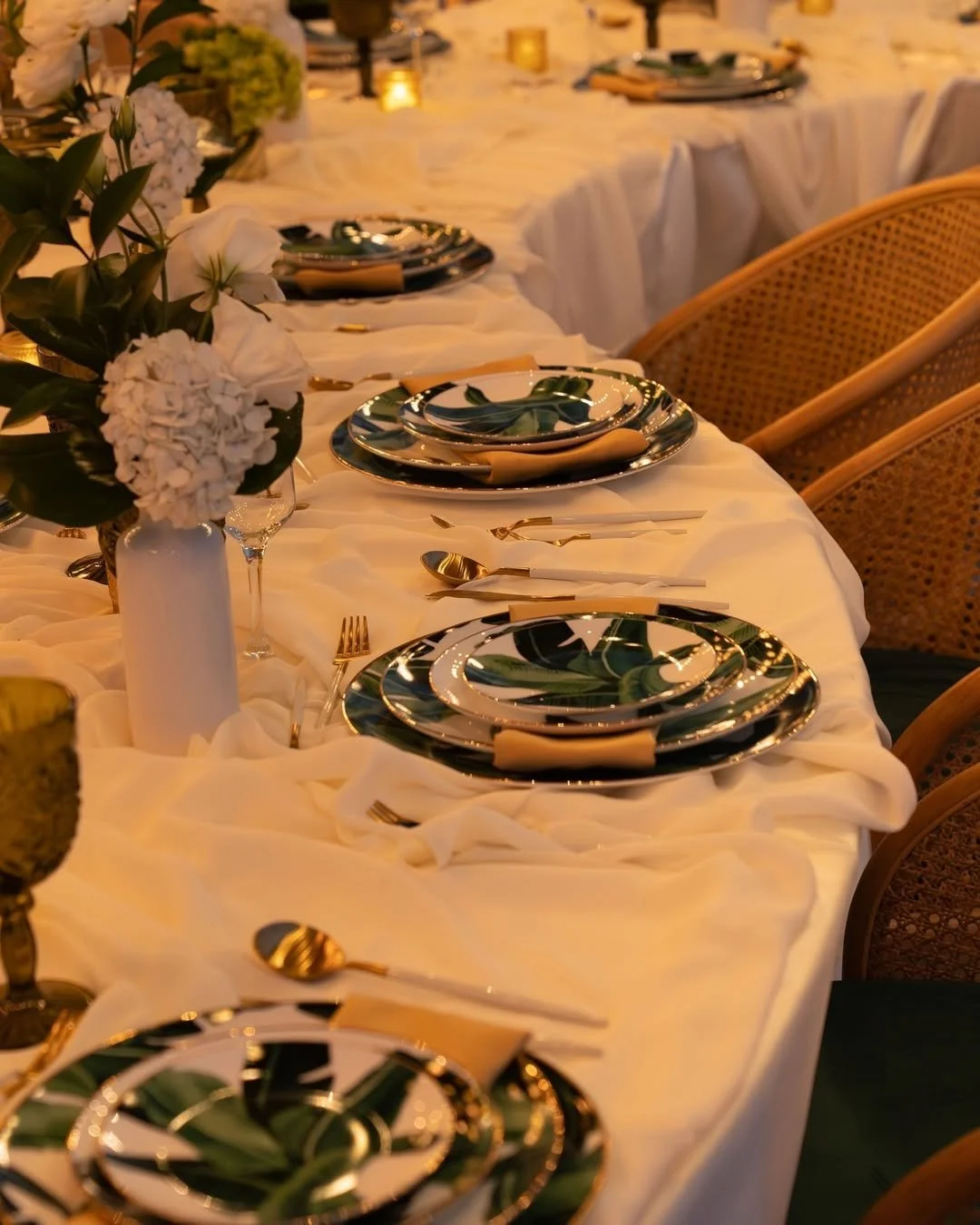 Mesa decorada con vajilla colorida, cubiertos dorados, jarrón con flores blancas y velas, en ambiente elegante para comida o evento especial.