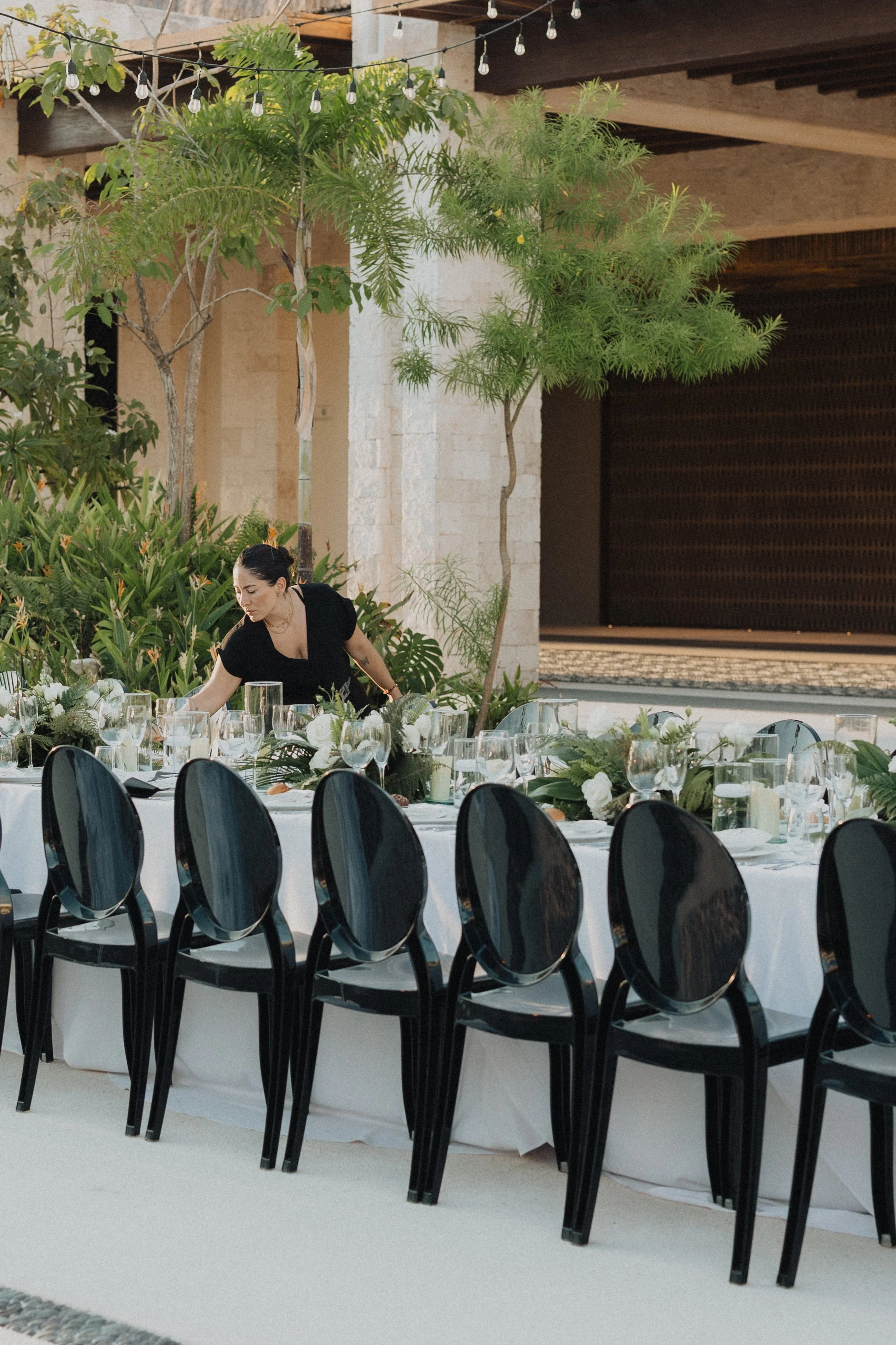 Mesa decorada con flores blancas, velas y cristalería en un evento al aire libre con silla negras y vegetación tropical en el fondo.