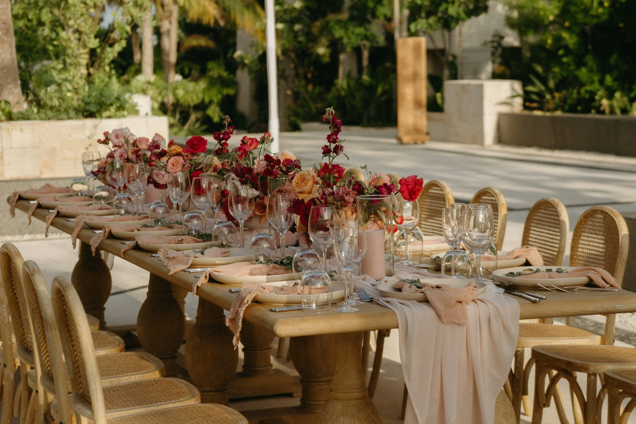Mesa decorada para evento al aire libre con flores, copas y platos, en entorno soleado con árboles y caminos.