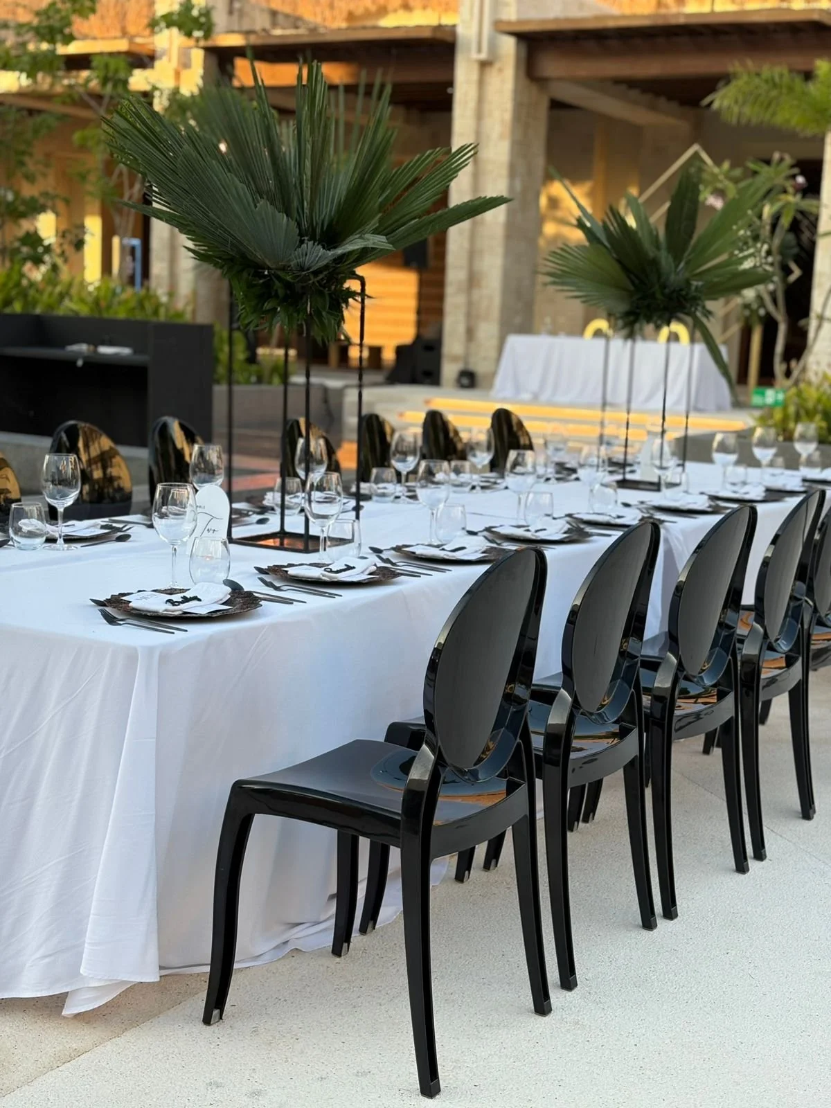 Mesa decorada con mantel blanco, vasos, cubiertos y centros de mesa con hojas grandes verdes en soporte negro, en un lugar al aire libre.