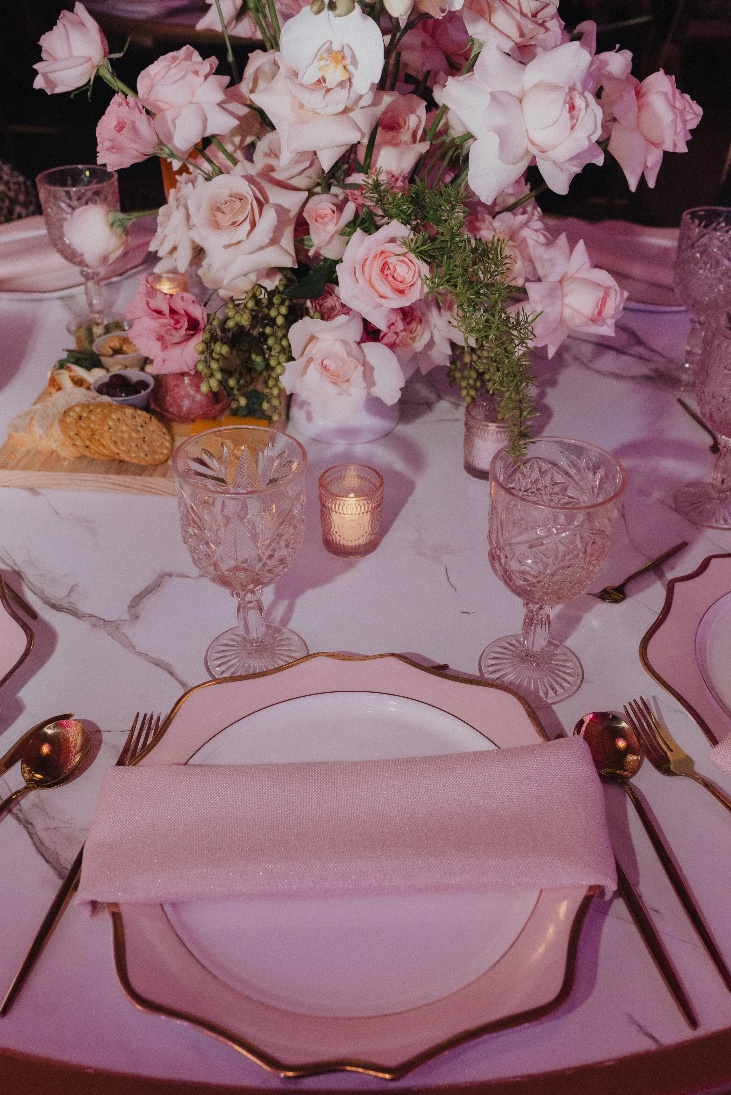 Mesa decorada con florero grande con rosas y otras flores rosadas, copas de cristal, platos de porcelana con borde dorado, manteles y servilletas rosadas, velas pequeñas. Se observa comida y utensilios de cena.
