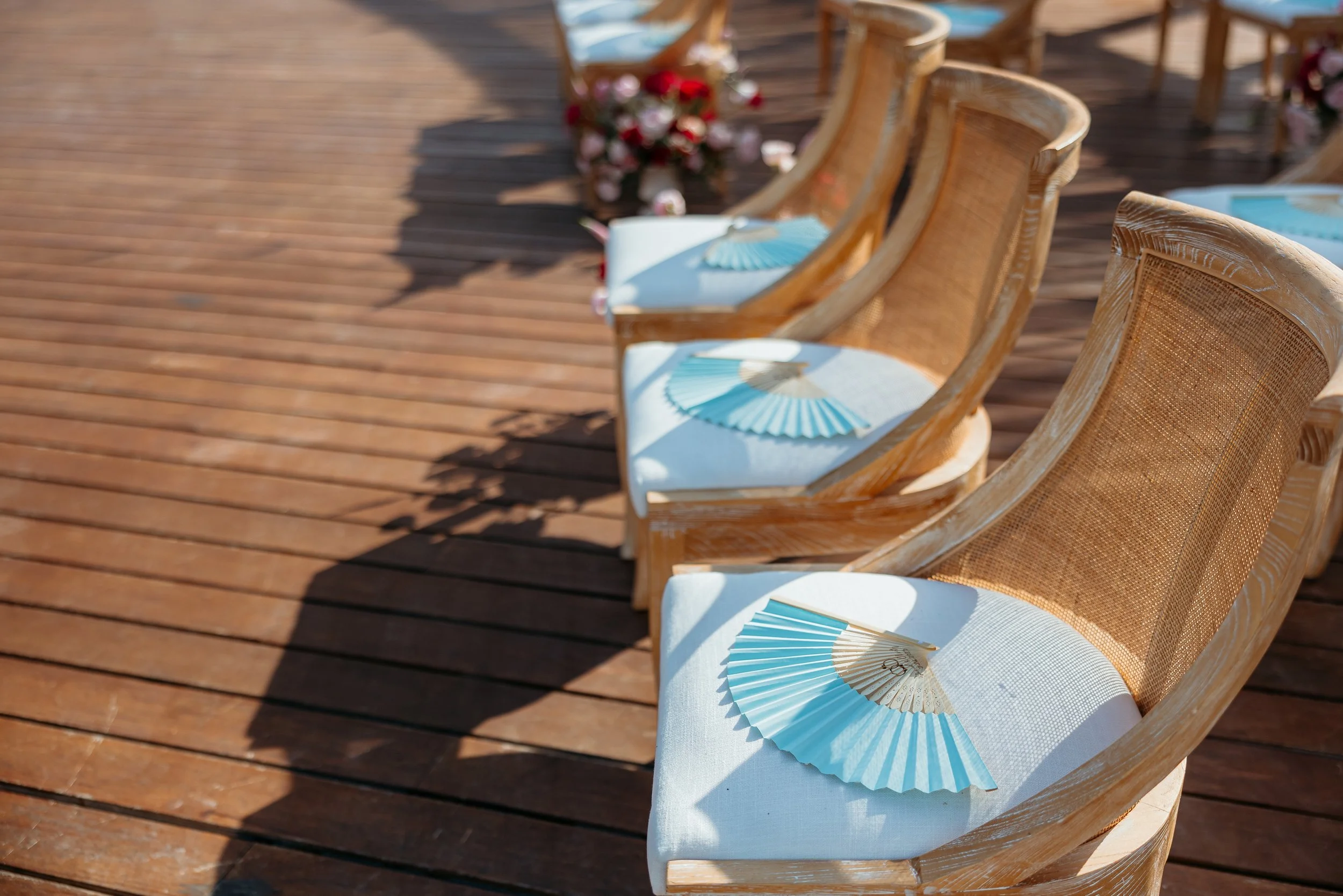 Sillas de madera con cojines blancos, decoradas con abanicos y arreglos florales en una terraza de madera.