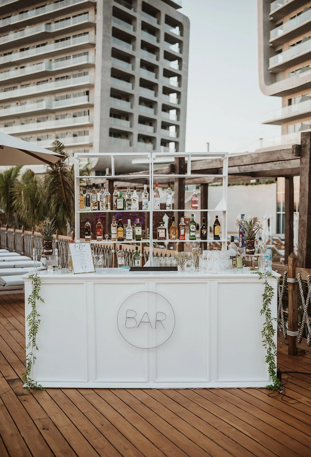 Bar blanco en una terraza con edificios modernos al fondo, decorado con plantas y sombrillas