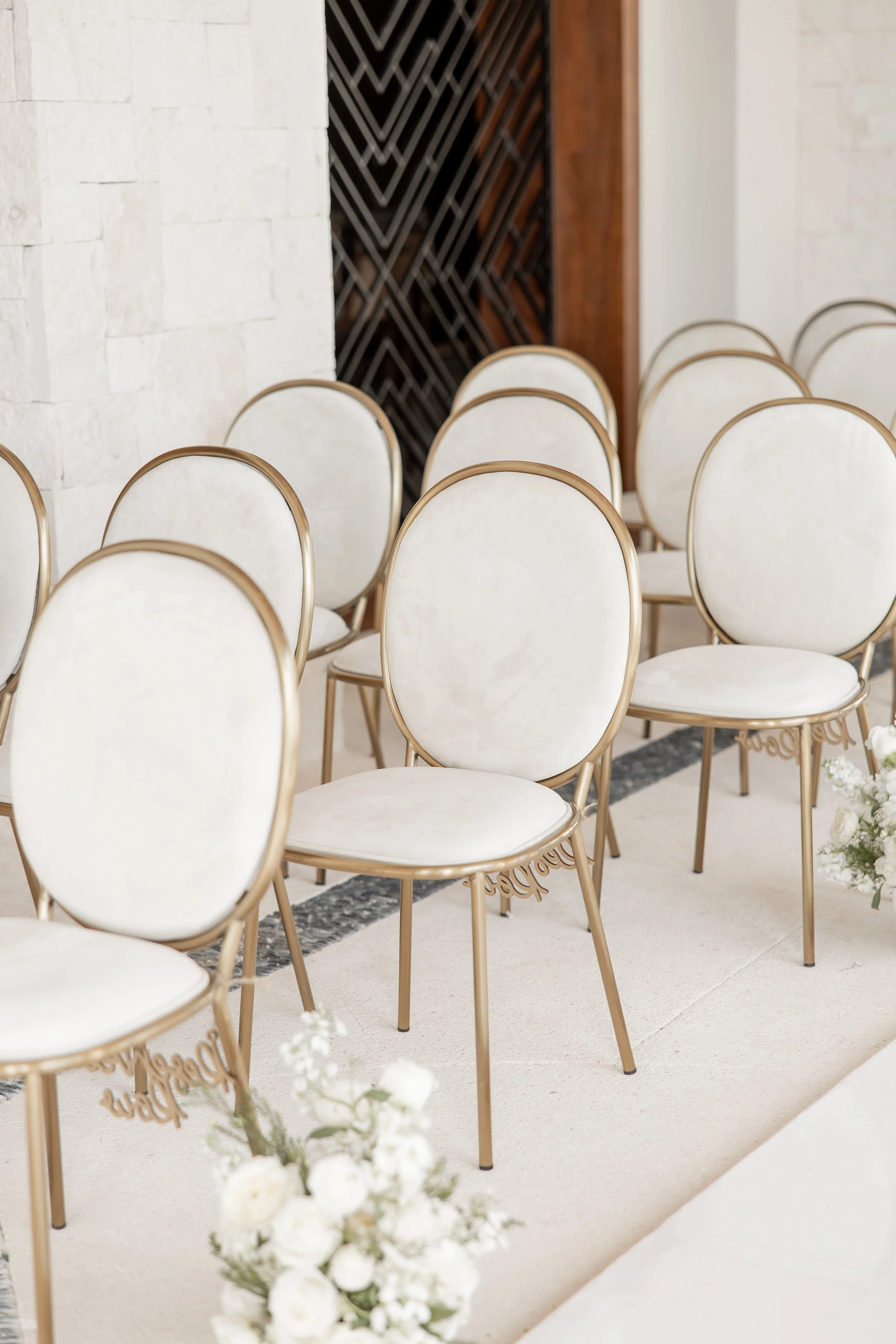 Sillas elegantes beige con marco dorado en una ceremonia, decoradas con flores blancas al frente.