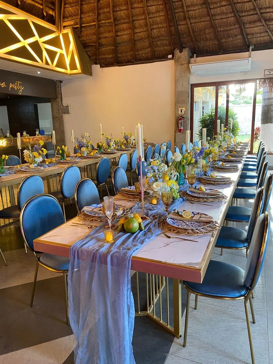 Mesa larga decorada con flores, velas, frutas y vajilla elegante en un espacio con decoración moderna y techo de paja.