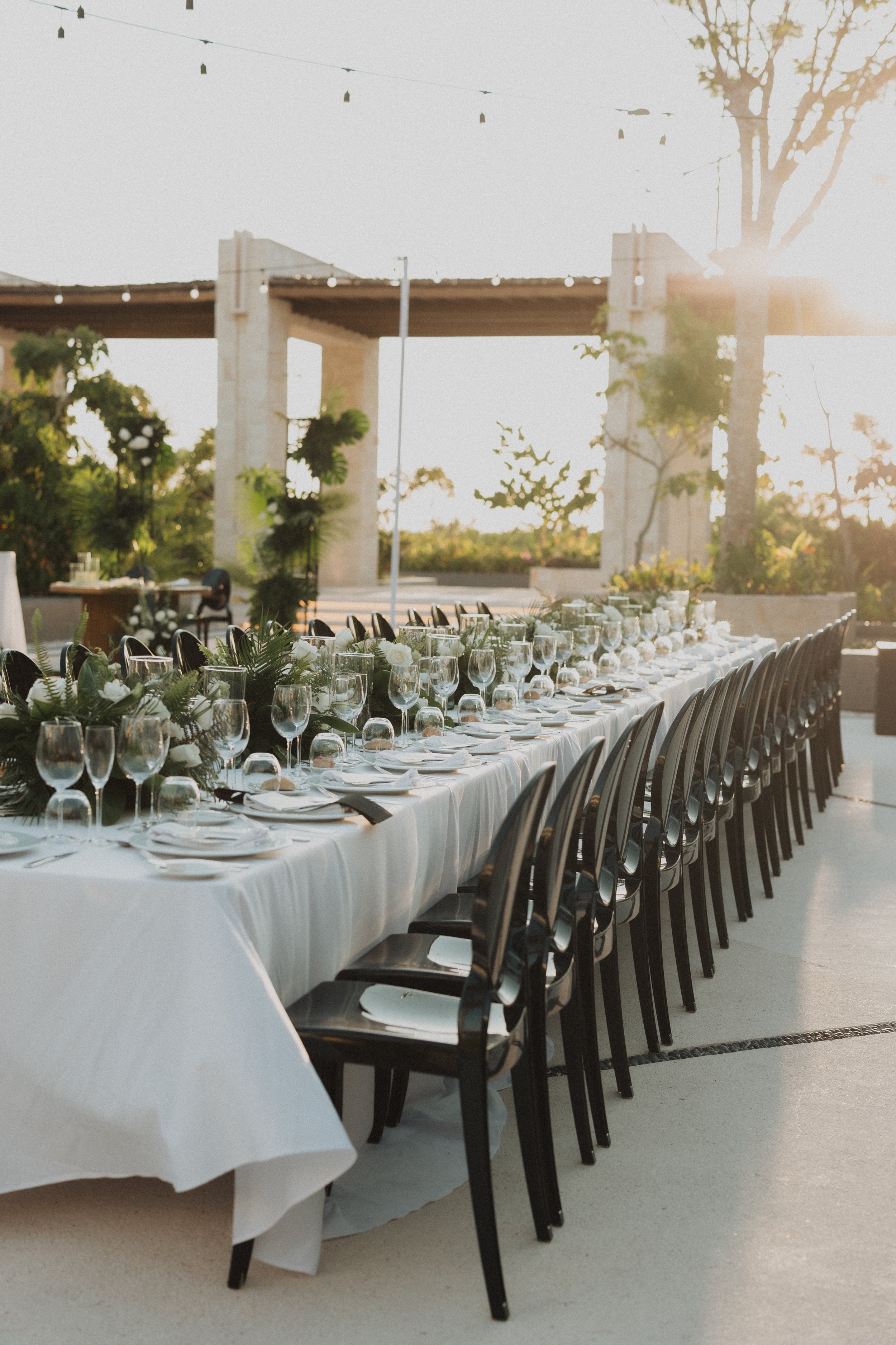 Mesa larga decorada con manteles blancos, vajilla y cristalería, rodeada de sillas negras, en un lugar al aire libre con árboles y una estructura de fondo, durante el atardecer