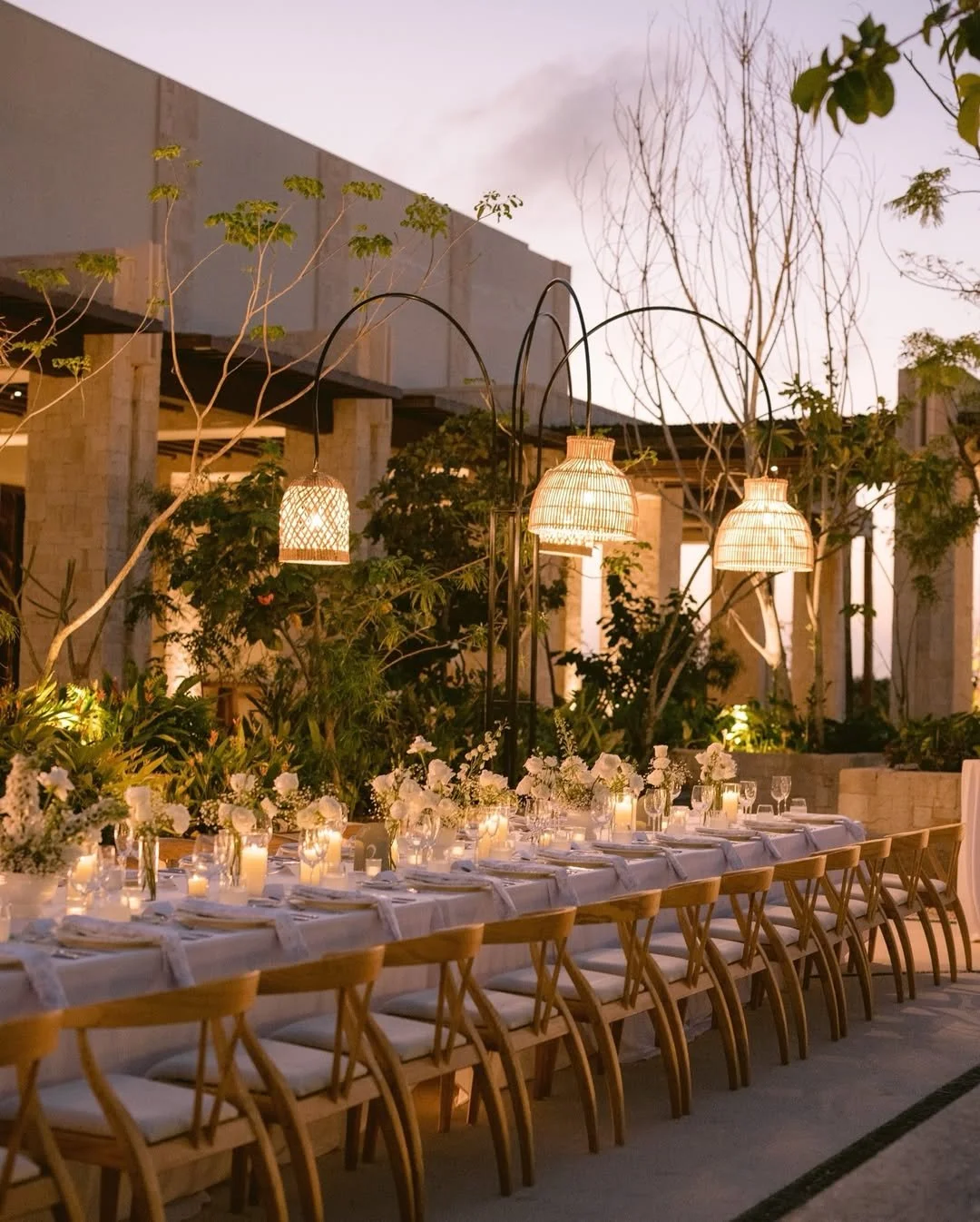 Mesa larga decorada con velas y flores, iluminada por lámparas de mimbre, en un ambiente exterior con plantas y árboles en la noche.