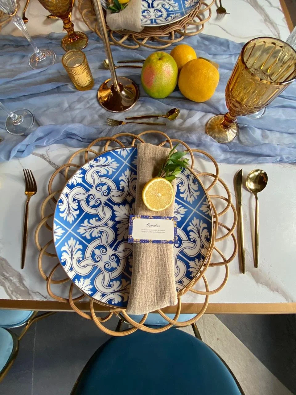Mesa decorada con platos azules y blancos, cubiertos dorados, copas de vino rosado y vasos de color ámbar, un centro de frutas con limón, manzana y cítricos, y ambiente elegante con detalles en dorado y azul.