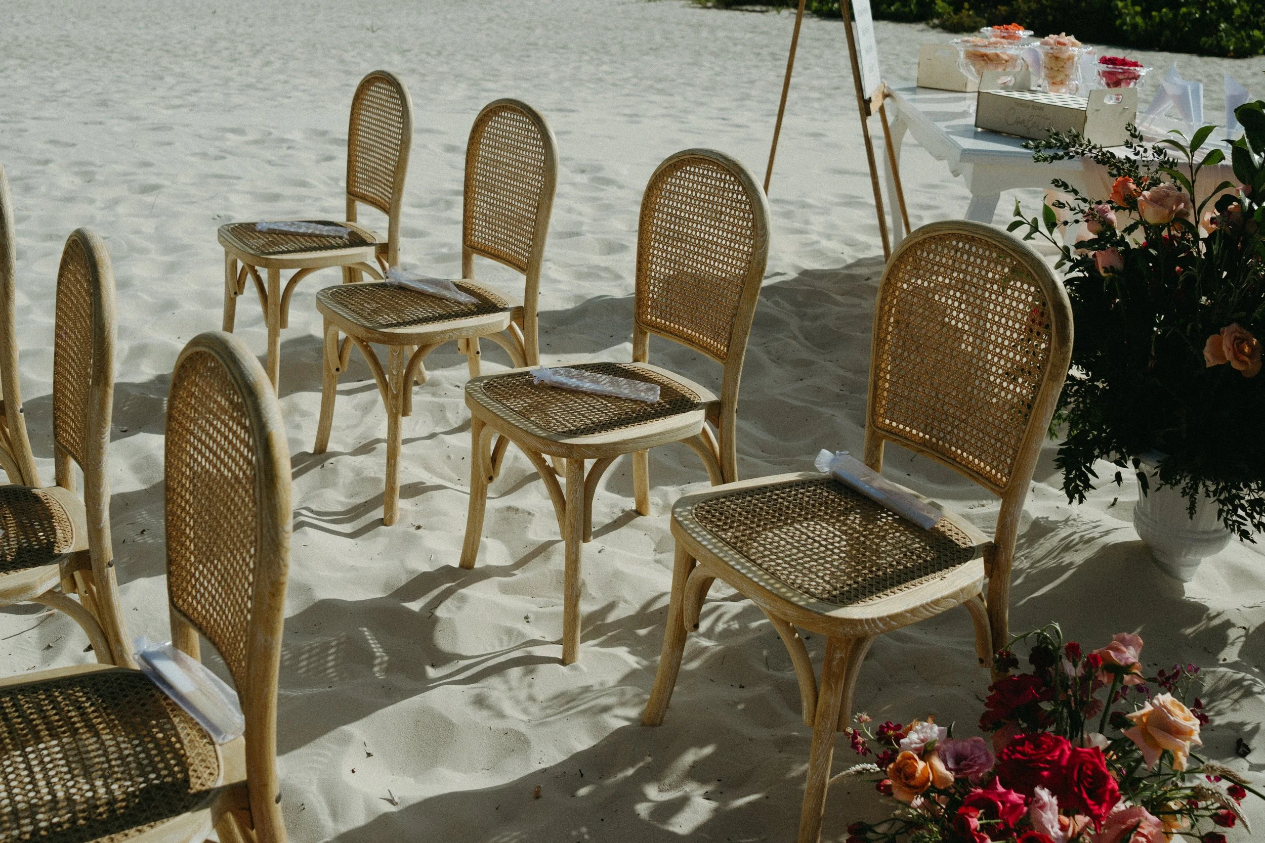 Sillas de mimbre en una ceremonia al aire libre en la arena, decoradas con flores y una mesa con dulces y otros objetos en el fondo.