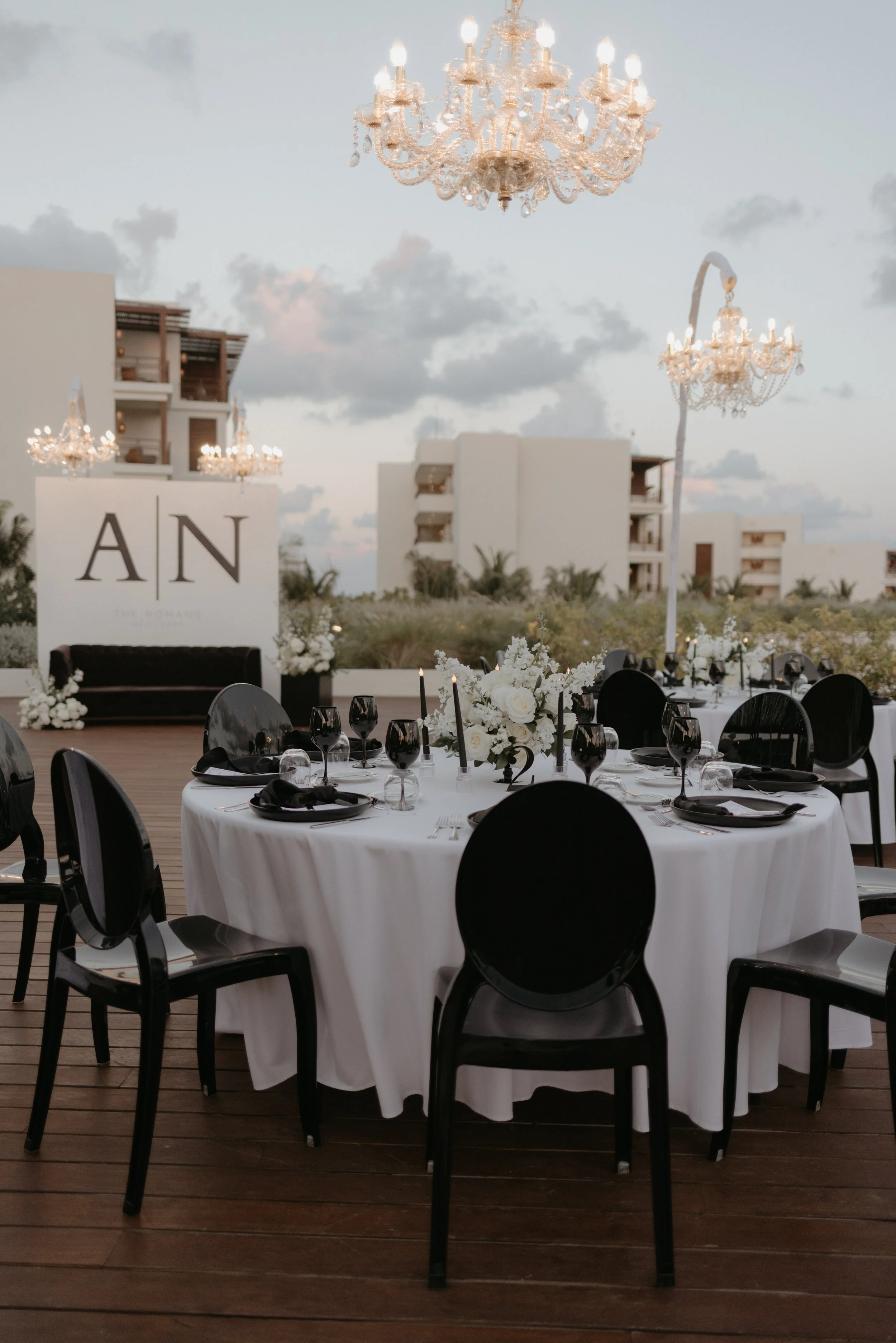 Una mesa elegante de cena al aire libre con manteles blancos, cubiertos plateados, copas negras, flores blancas y candelabros, decorada para un evento en la tarde, con edificios modernos y lámparas de araña en el fondo.