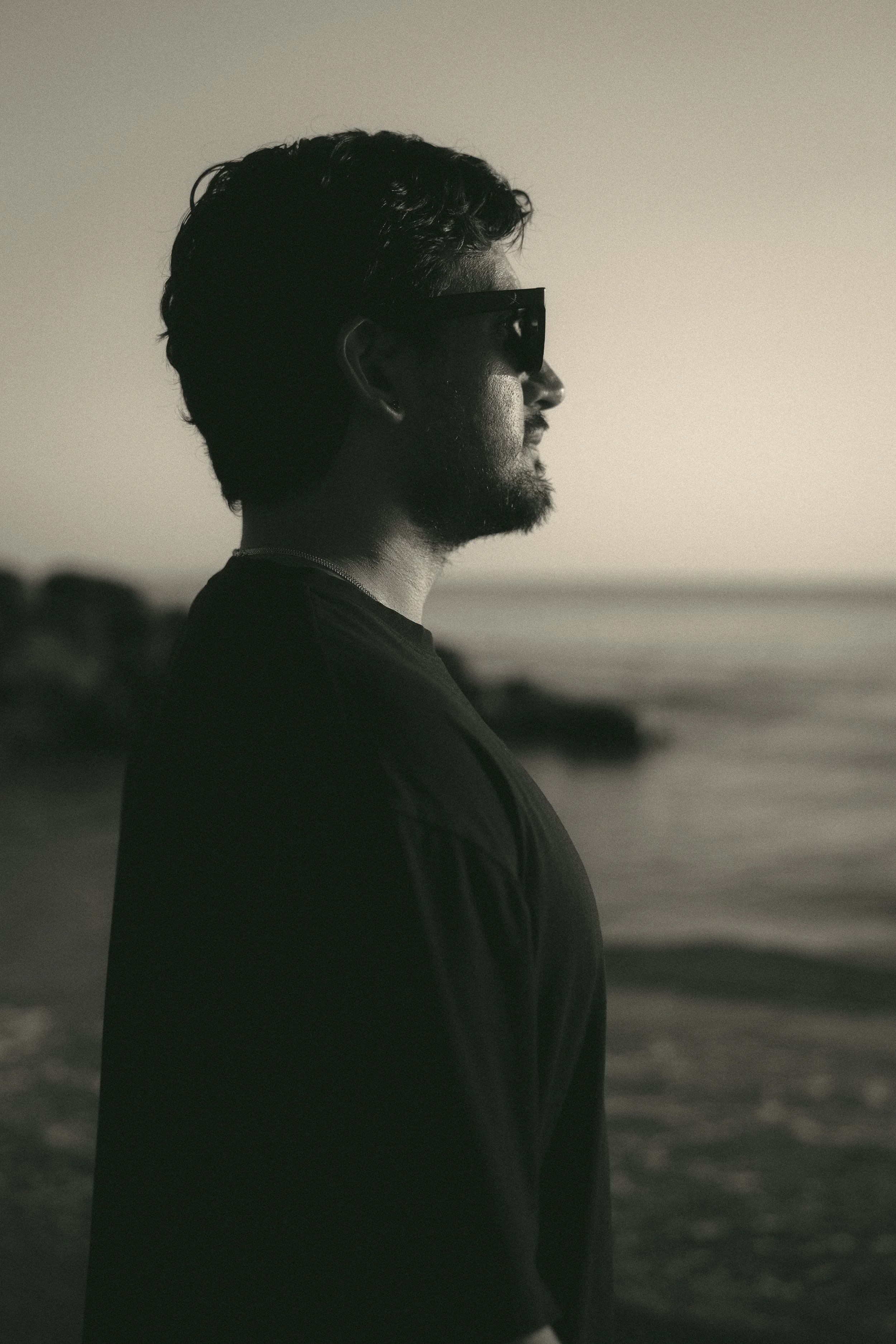 Side profile of a man with sunglasses standing on the beach during sunset or sunrise, with ocean and blurred rocks in the background.