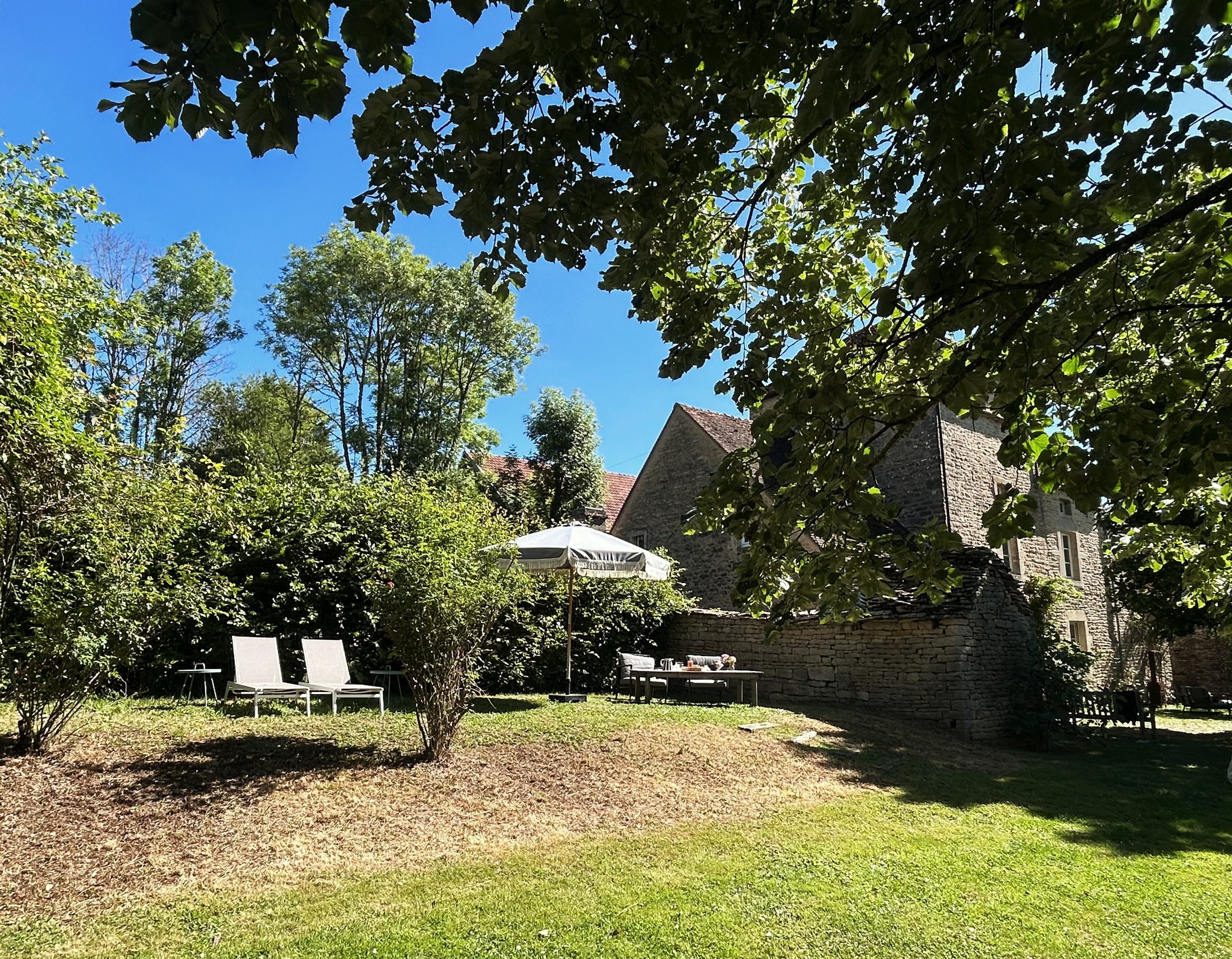 Jardin avec chaises longues et parasol sous un arbre, maison en pierre en arrière-plan, ciel clair.