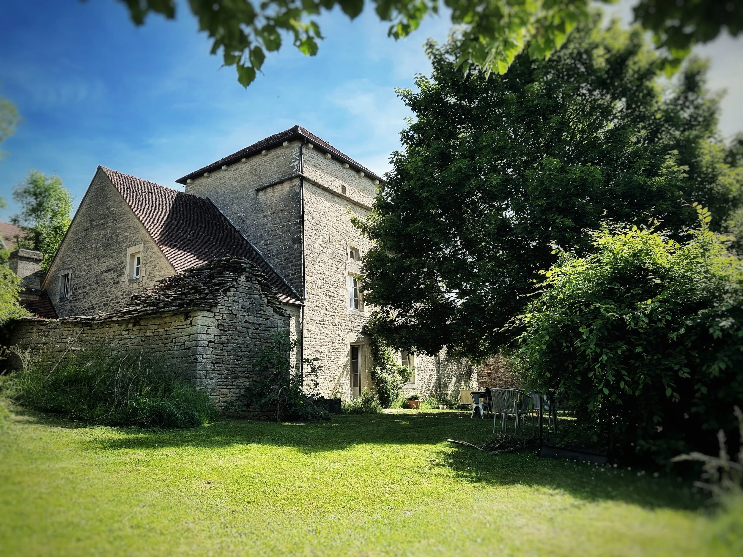 Une ancienne maison en pierre avec un toit en tuiles, entourée d'arbres verts, avec un jardin et une zone de détente avec des chaises.