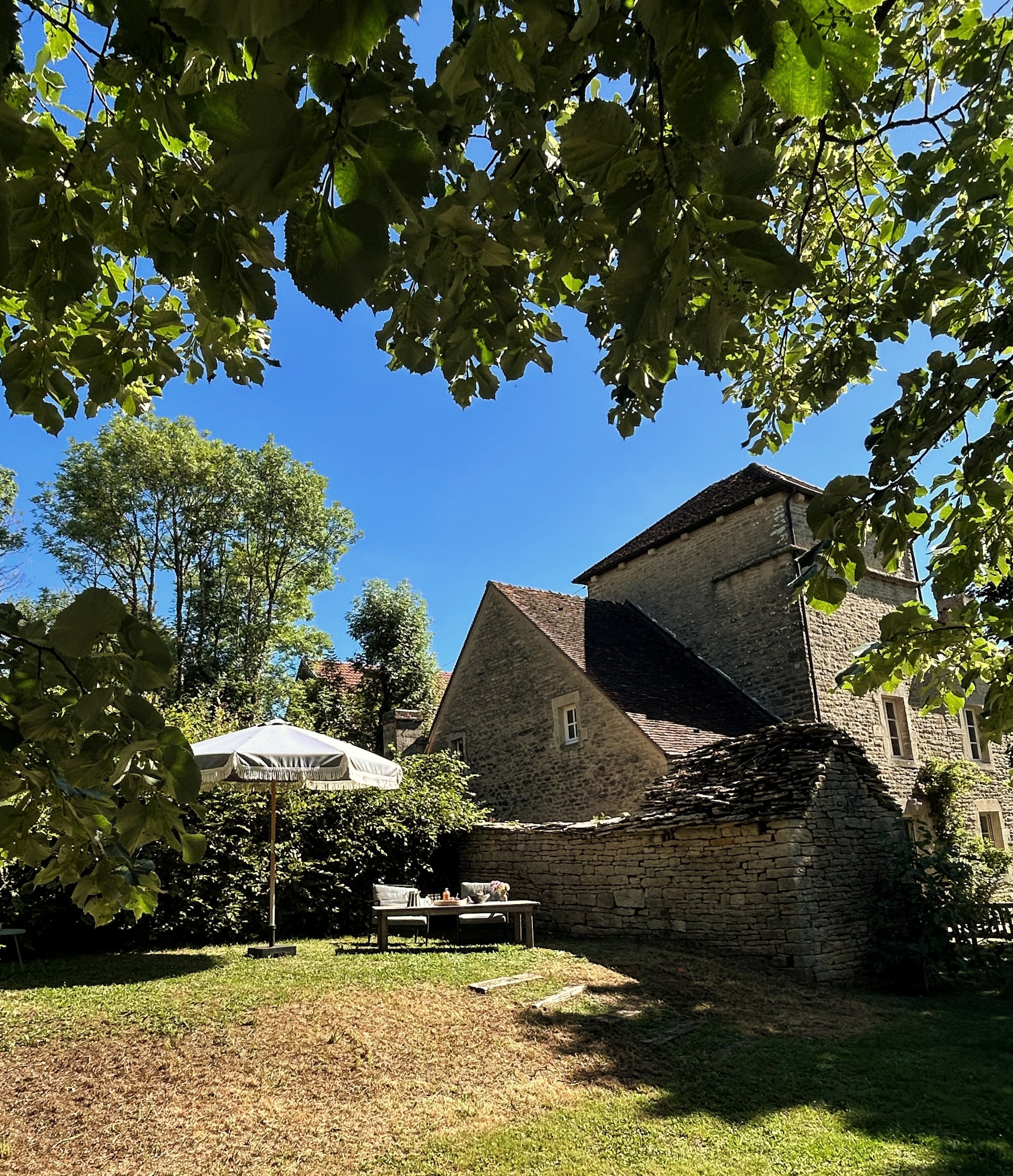 Jardin dans une cour avec un parasol blanc, une table et des chaises, entouré de murs en pierre et d'arbres feuillus, sous un ciel bleu.
