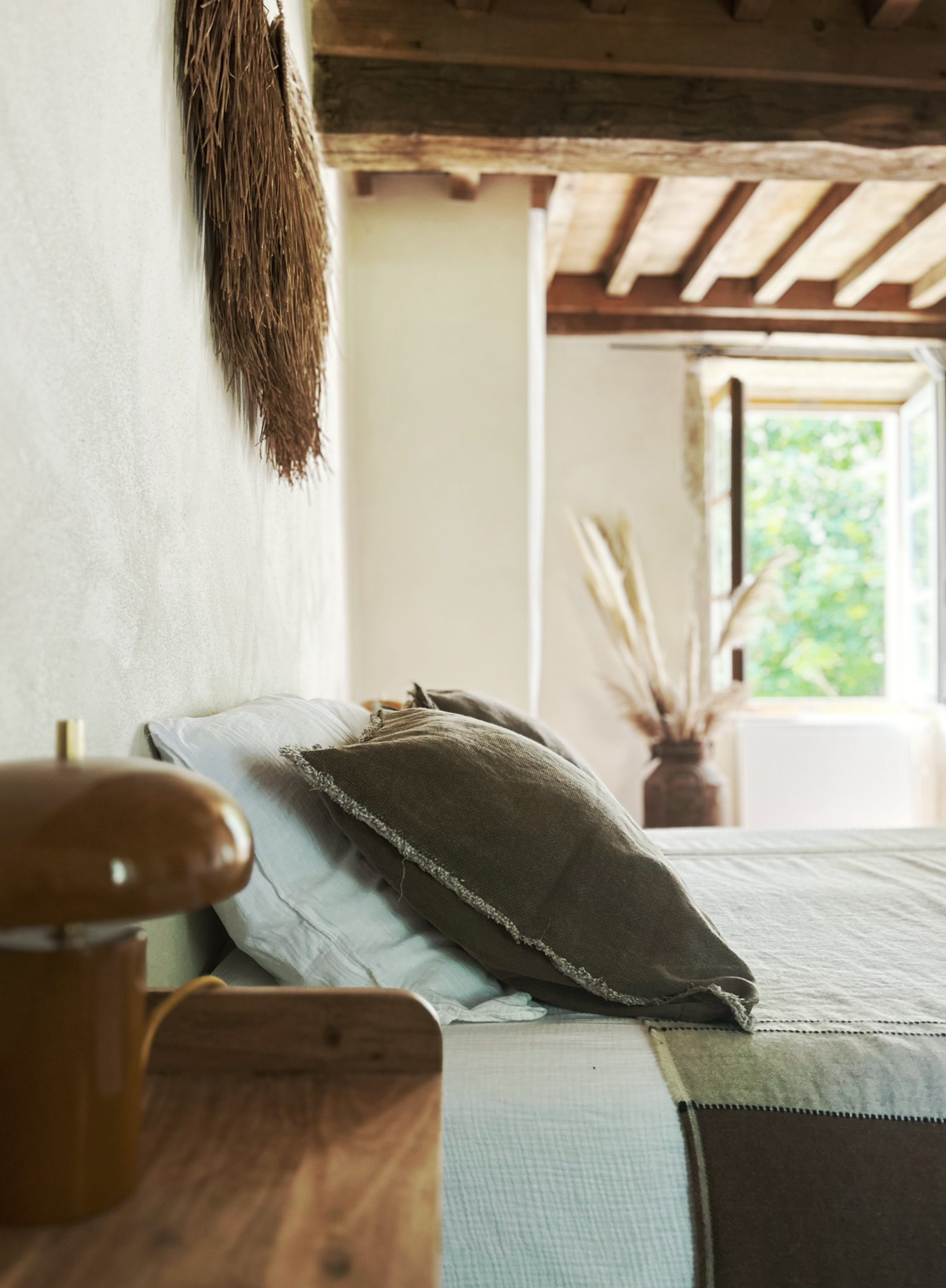 Intérieur d'une chambre avec un lit fait, plusieurs coussins, une fenêtre laissant entrer la lumière naturelle, et un plafond en bois avec poutres apparentes.