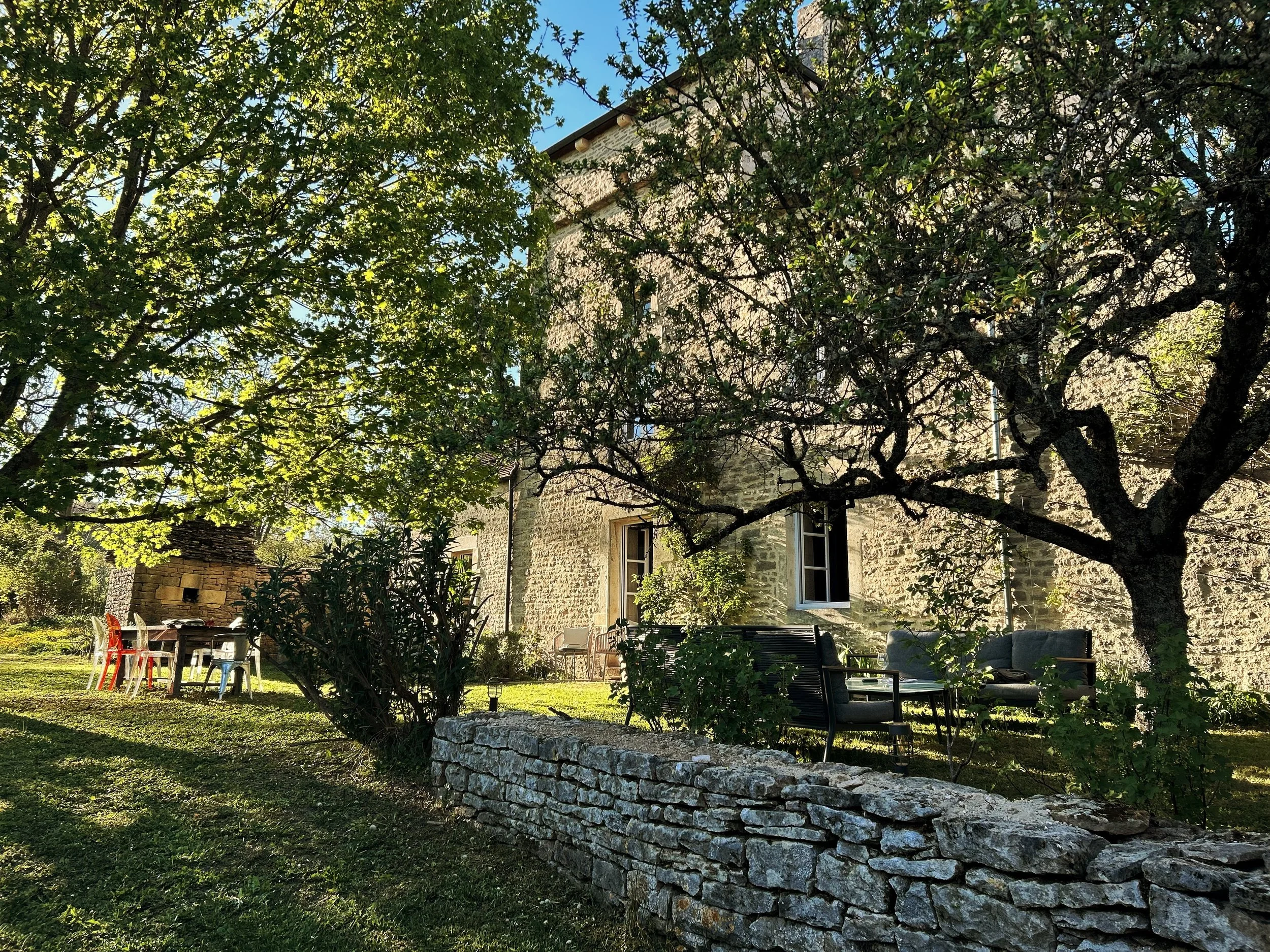 Jardin avec arbres feuillus, terrasse avec des chaises colorées, une maison en pierre et un mur en pierre, mobilier de jardin, ambiance ensoleillée.