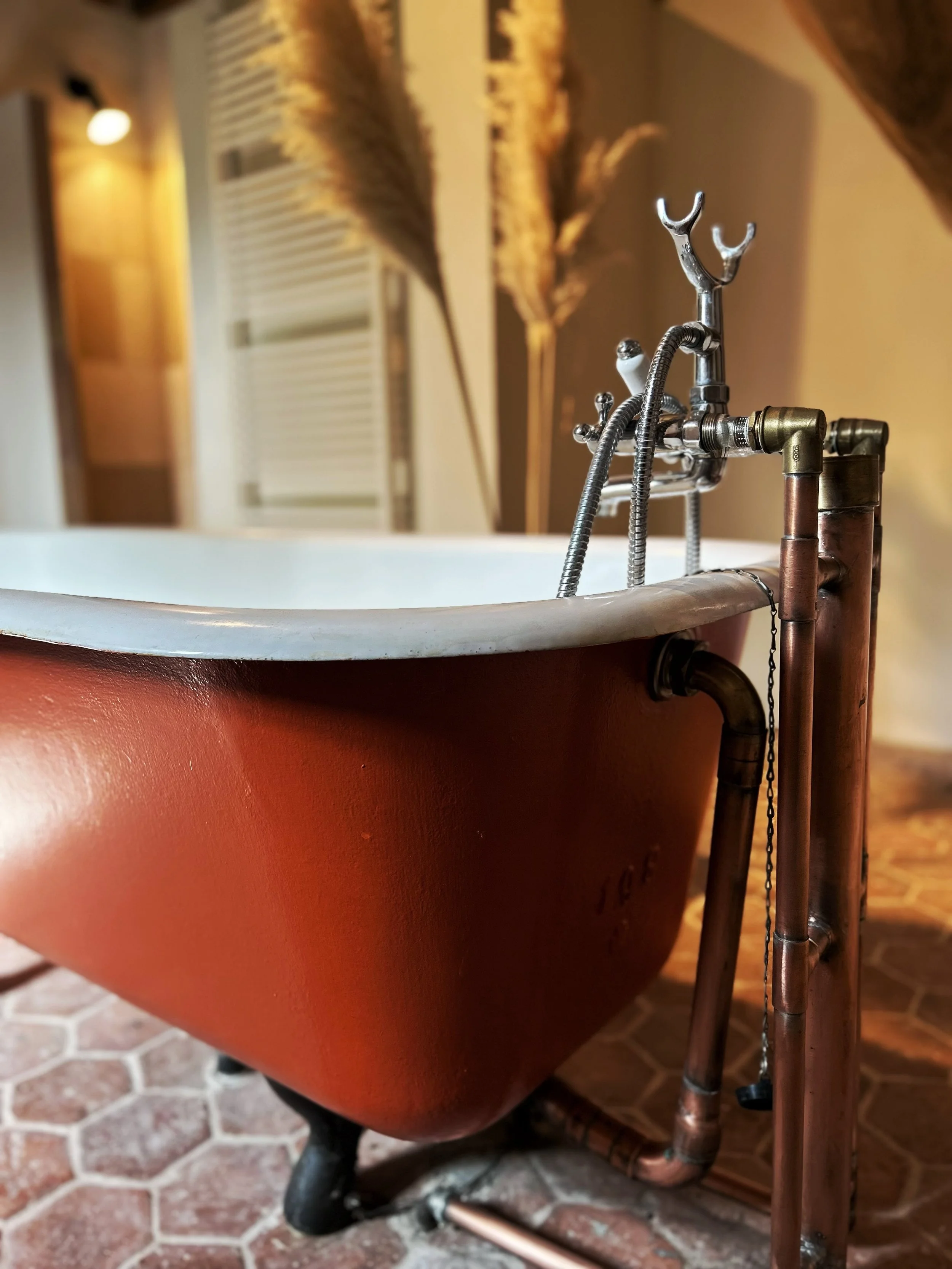 Une baignoire ancienne en cuivre avec une robinetterie fait main en métal, entourée de plantes séchées et un sol en carreaux hexagonaux.