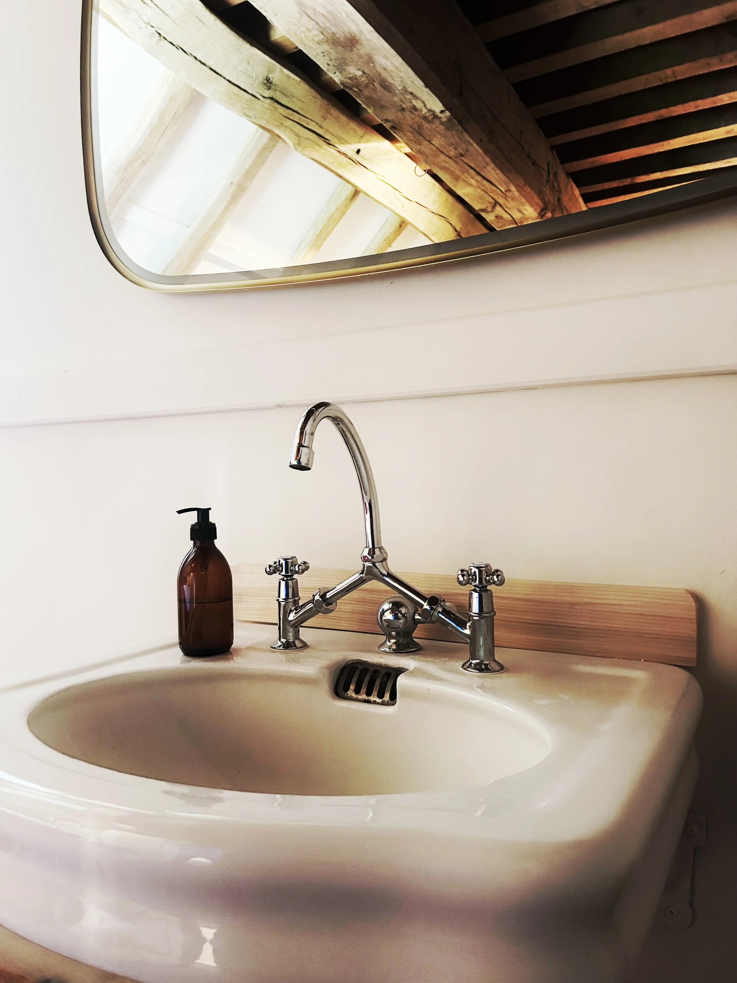Lavabo blanc avec robinet en métal, sur un meuble en bois clair, avec un miroir au mur et une bouteille de savon liquide marron à gauche, le tout dans une salle de bain avec un plafond en bois apparent.
