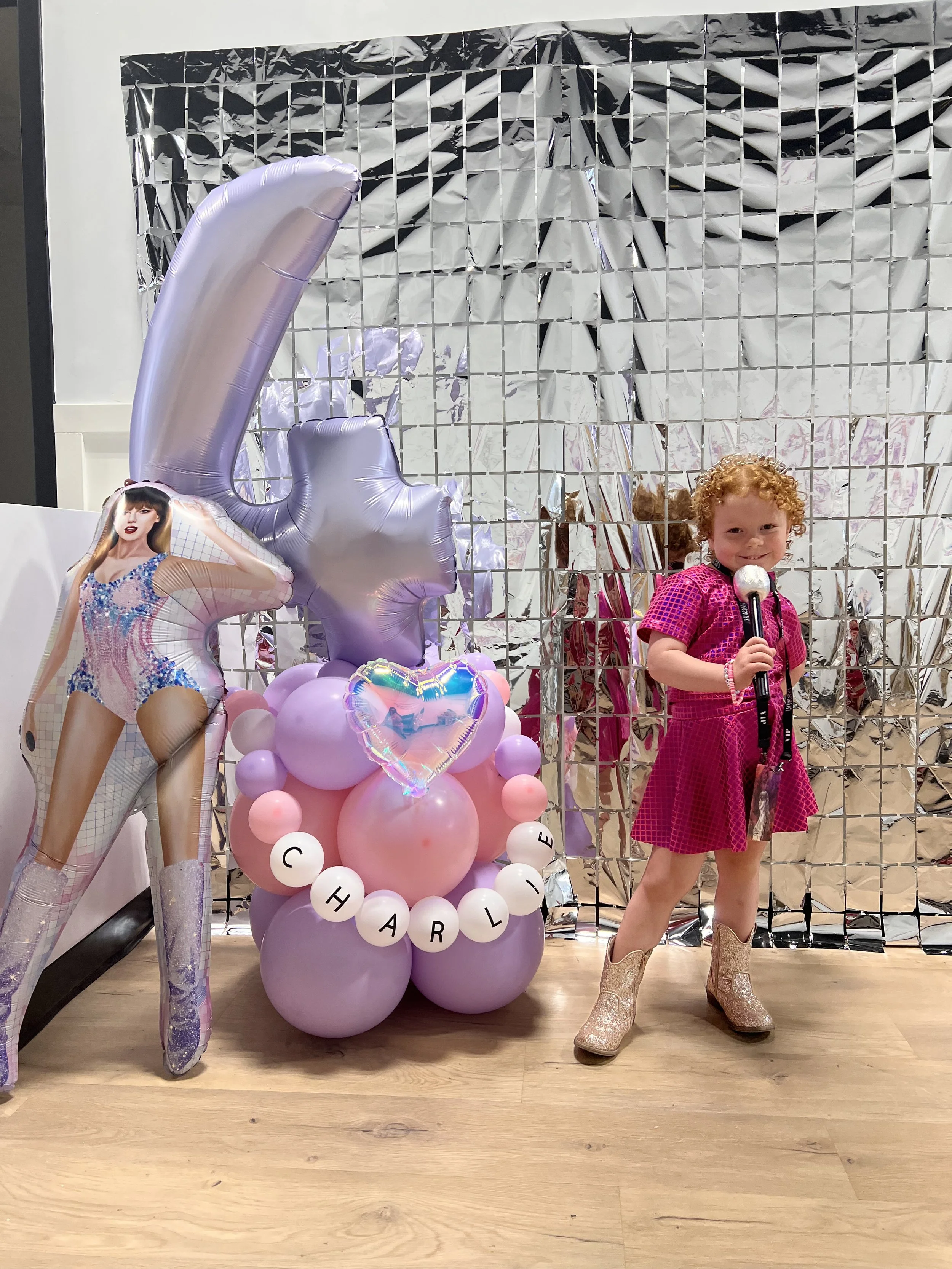 holds a microphone and stands in front of a shiny silver backdrop with balloons. The balloons include a large purple number '1', pink, purple, and white balloons, and a heart-shaped balloon with an iridescent finish. The balloons spell out 'Charlye'