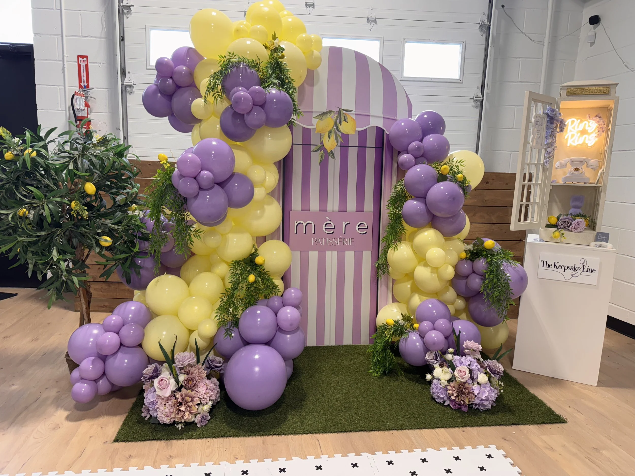 Decorative balloon arrangement and floral display at a pastel-colored event for 'mère pâtisserie', with a striped purple and white backdrop and greenery, on a green grass-like mat, inside a well-lit room.