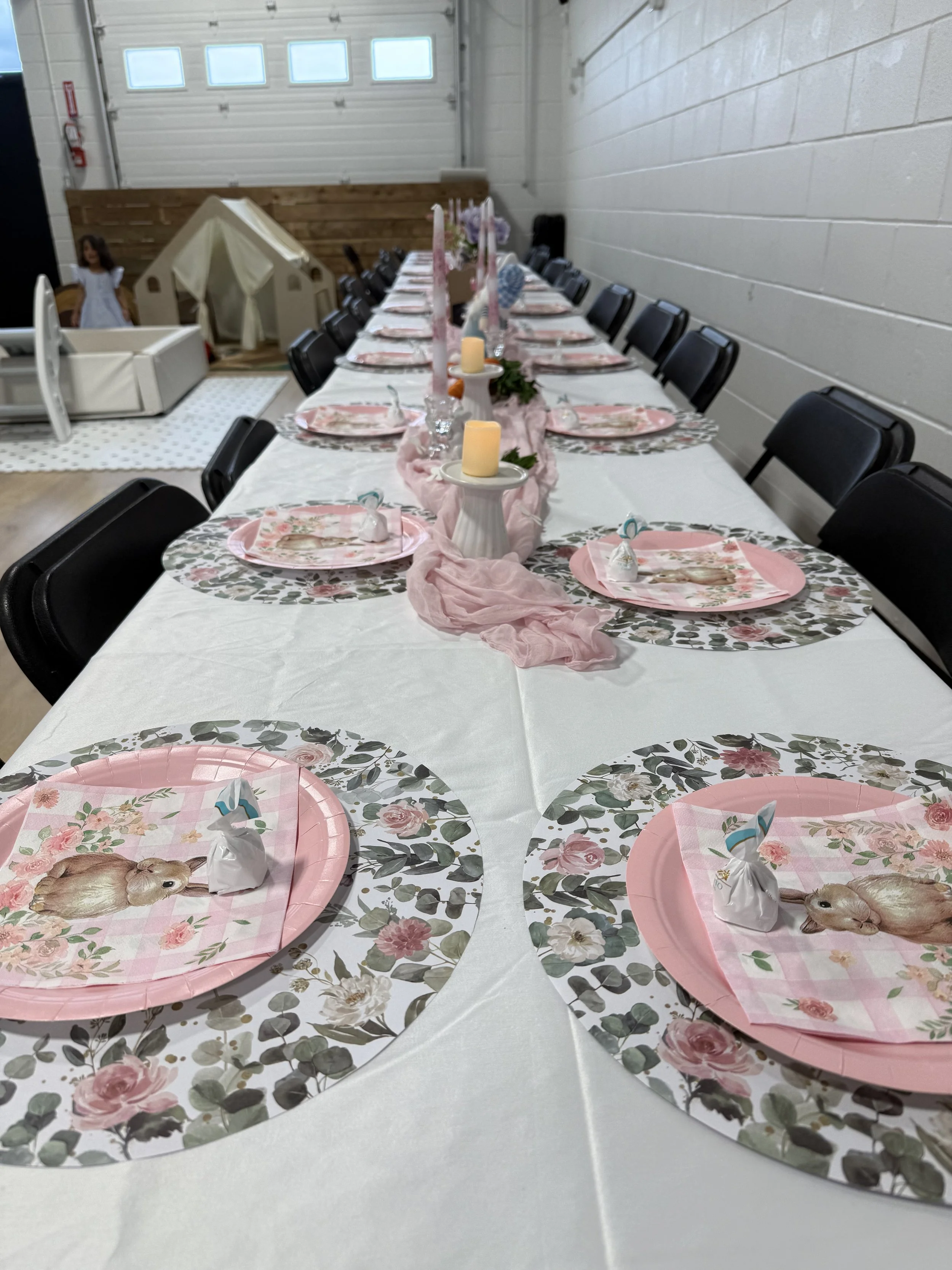 Long table decorated for a tea party or celebration, with floral placemats, pink trays, wrapped favors, candles, and a pink fabric runner, set in a room with a garage door and a play tent in the background.