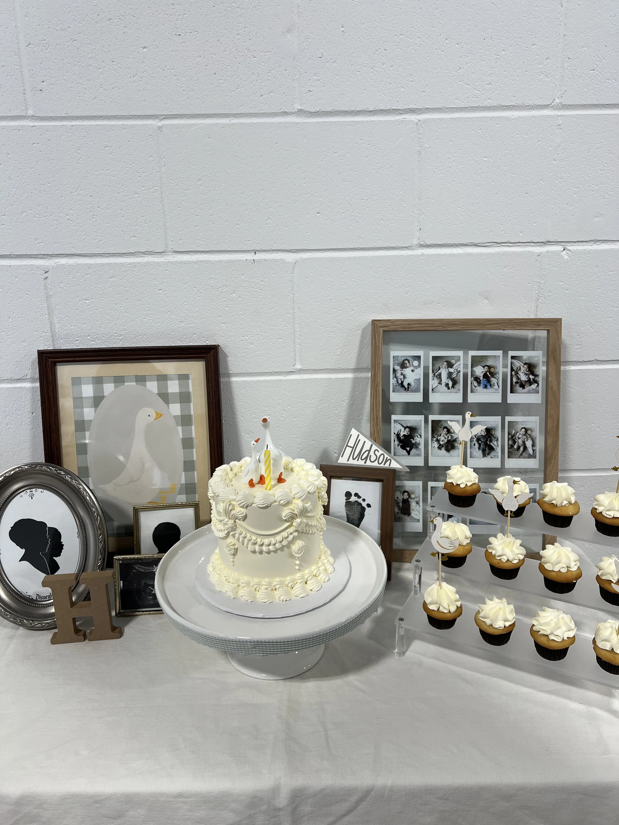 A decorated table with a white cake, cupcakes, framed pictures, and a sign that says 'Hudson' in a room with a white brick wall.