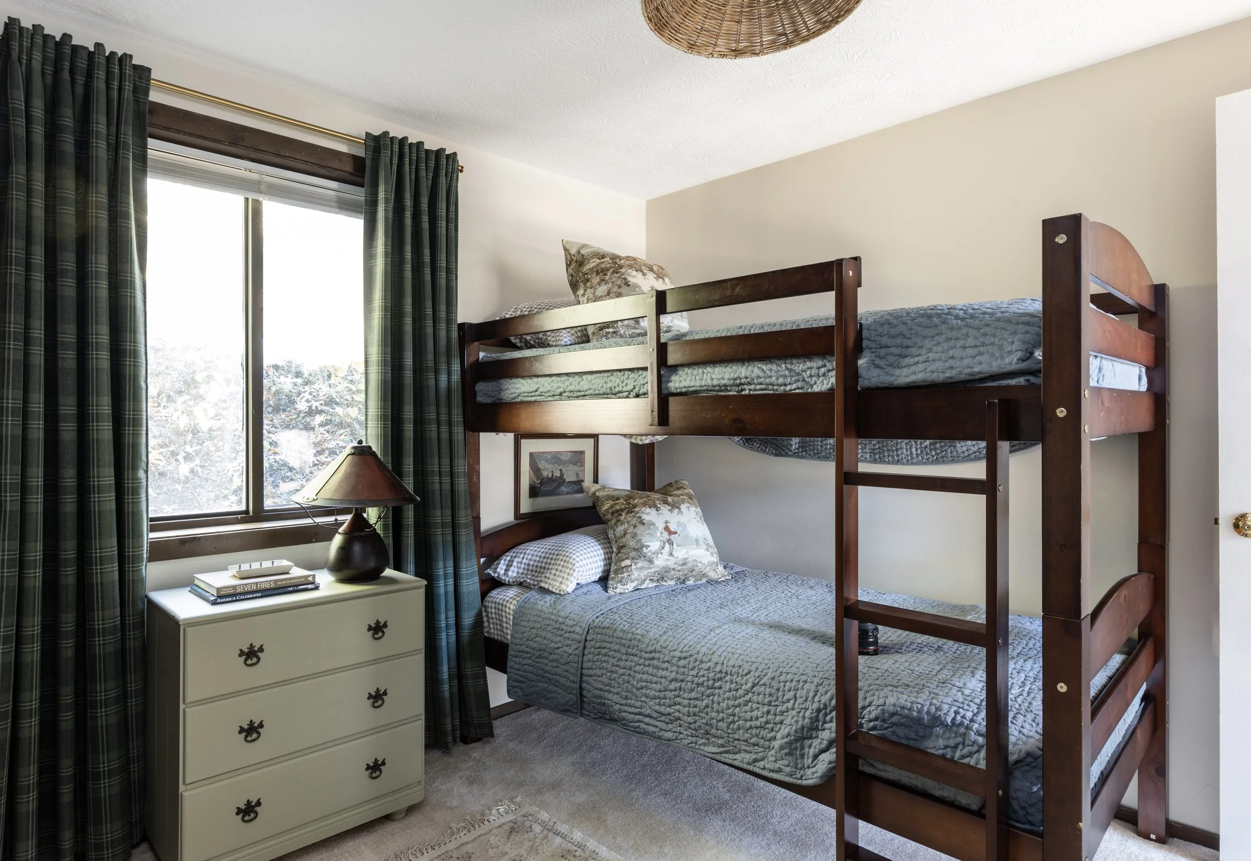 A bedroom with a bunk bed, a window with green plaid curtains, a white nightstand with books and a lamp, and framed pictures on the wall.