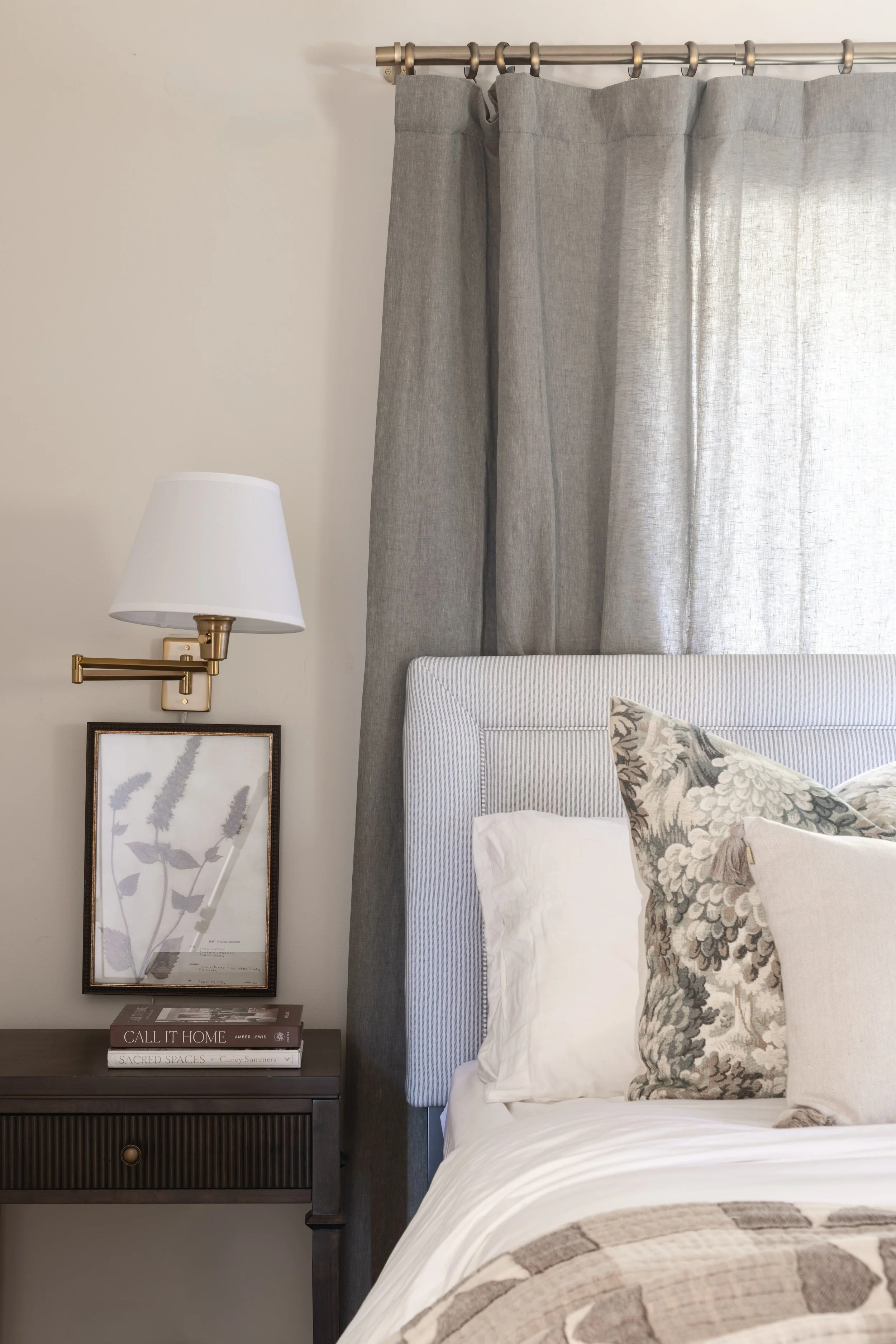 A bedroom corner with a bed, gray curtains, a wall-mounted lamp, a framed botanical print, and a nightstand with books.