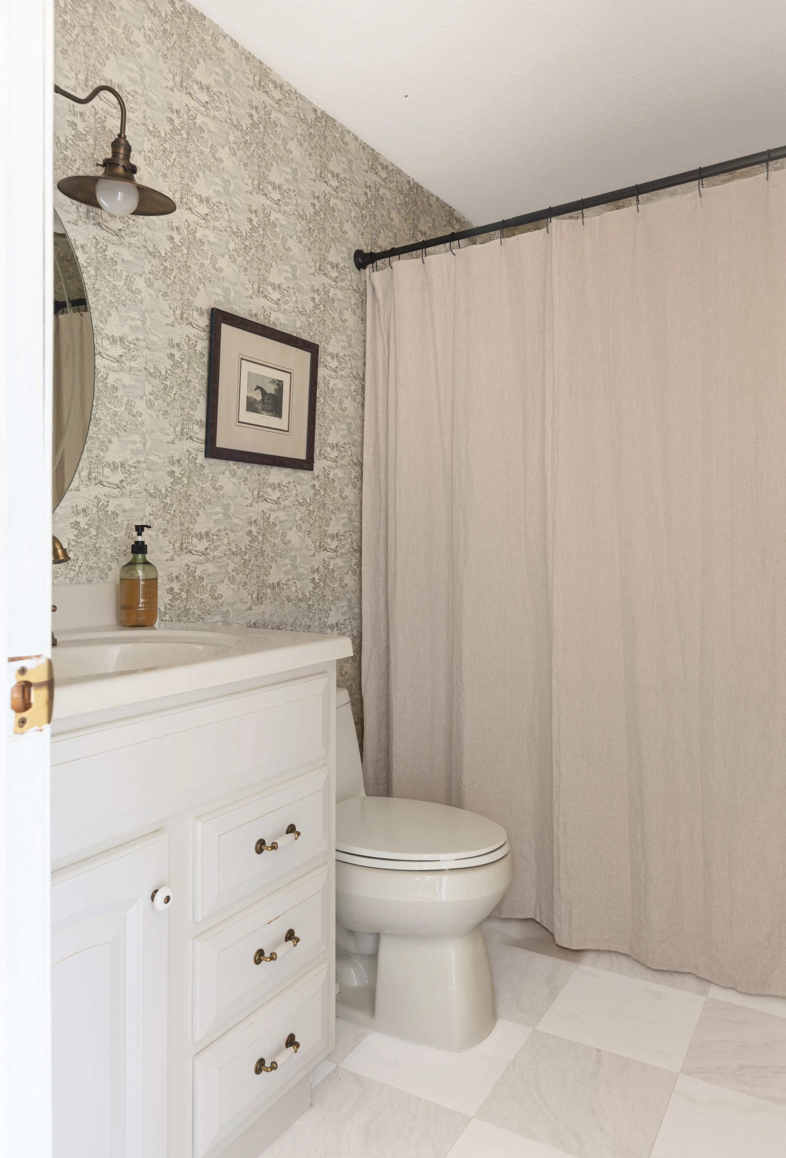 Small bathroom with a white vanity, dark bronze hardware, a framed picture of a horse, a beige shower curtain, a cream-colored wall, a brass sconce light fixture, a soap dispenser, and a toilet.