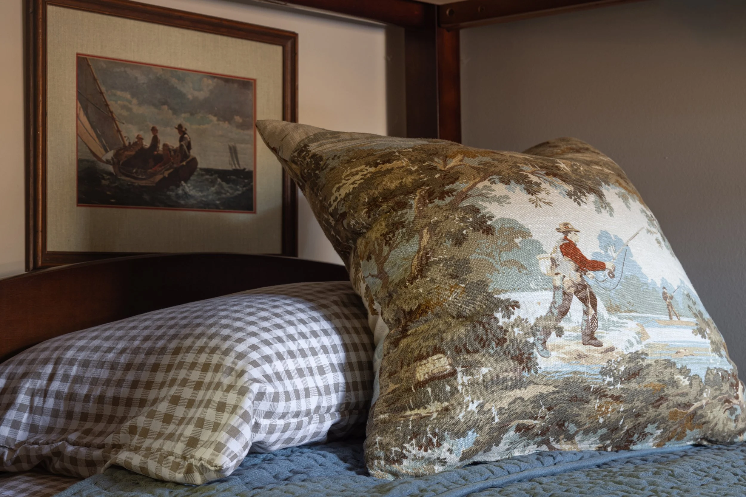 A close-up of a bed with checkered sheets, a decorative pillow featuring a person fishing in a wooded scene, and a framed painting of three children on a sailboat hanging on the wall.