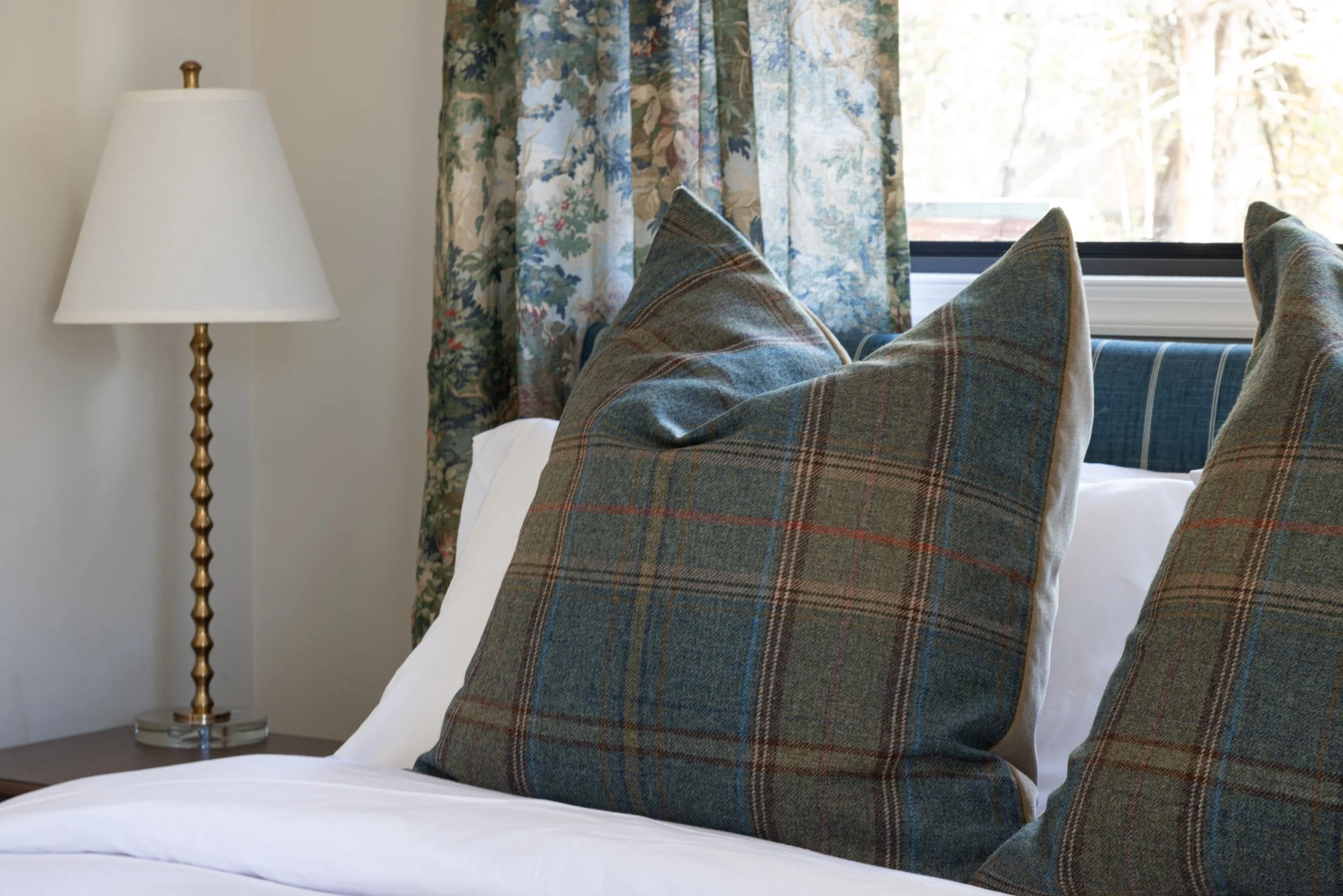 Close-up of a bed with plaid pillows, a bedside table with a gold lamp, floral curtains, and a window showing trees outside in a bedroom.