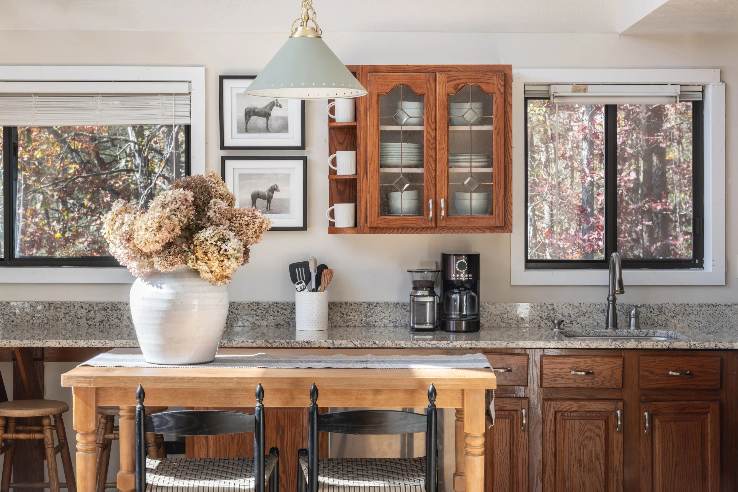 Kitchen with wooden cabinets, two windows, a table with a flower arrangement, coffee maker, and utensils.