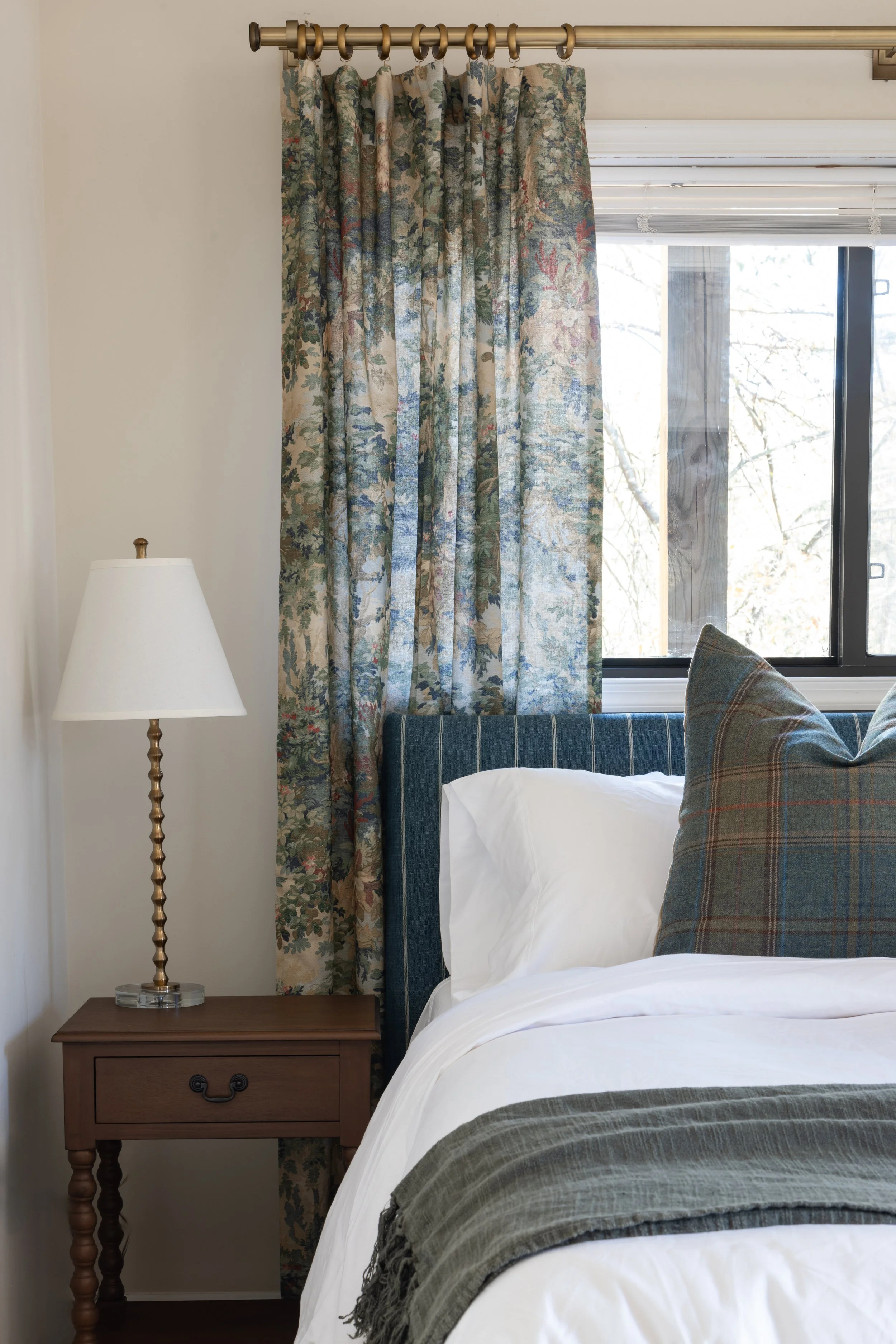 A bedroom corner with a wooden nightstand, white bed linens, a plaid pillow, a table lamp, a window with floral curtains, and a paneled window shade.