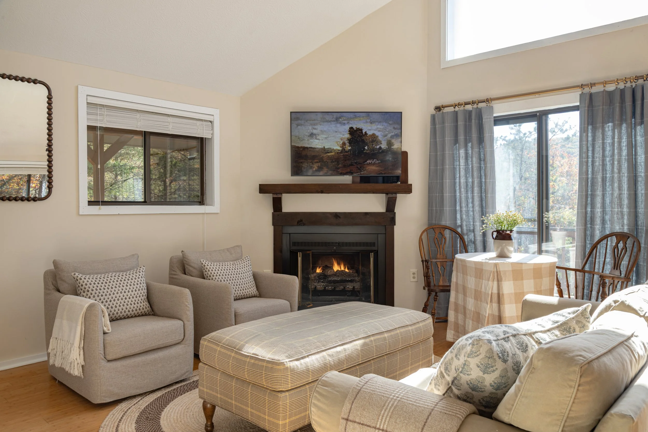 A cozy living room with a fireplace, a beige sofa with patterned cushions, a plaid ottoman, a wooden dining table with chairs, and large windows with curtains letting in natural light.