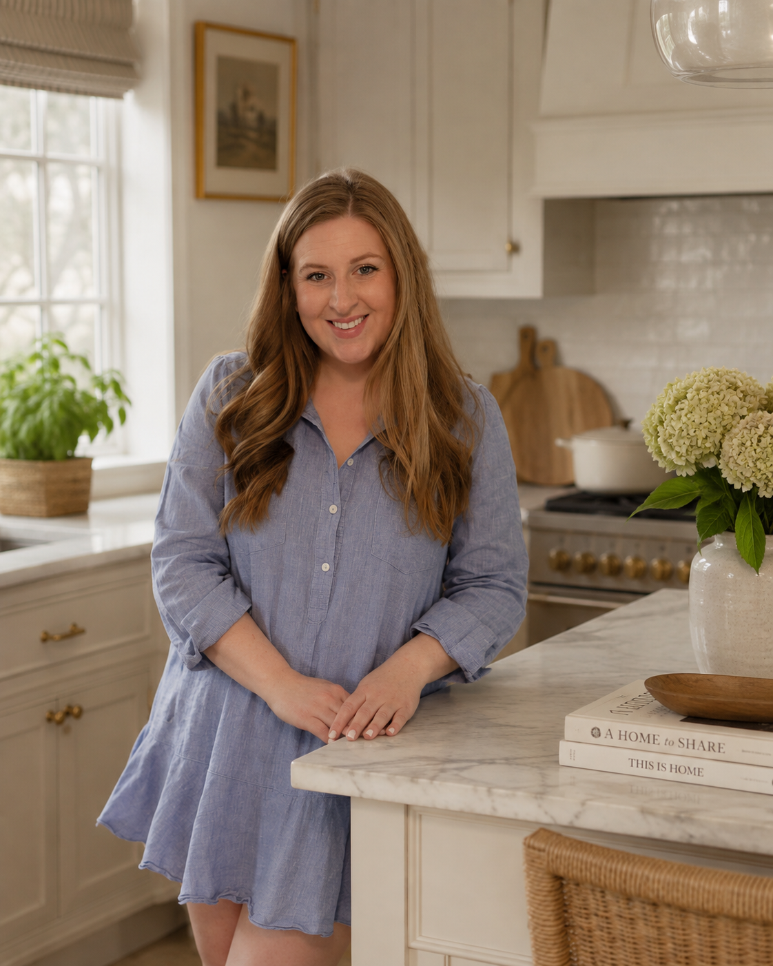 an interior designer with long red hair smiling in a bright kitchen, wearing a light blue dress, standing by a marble countertop with books and a vase of white hydrangeas.