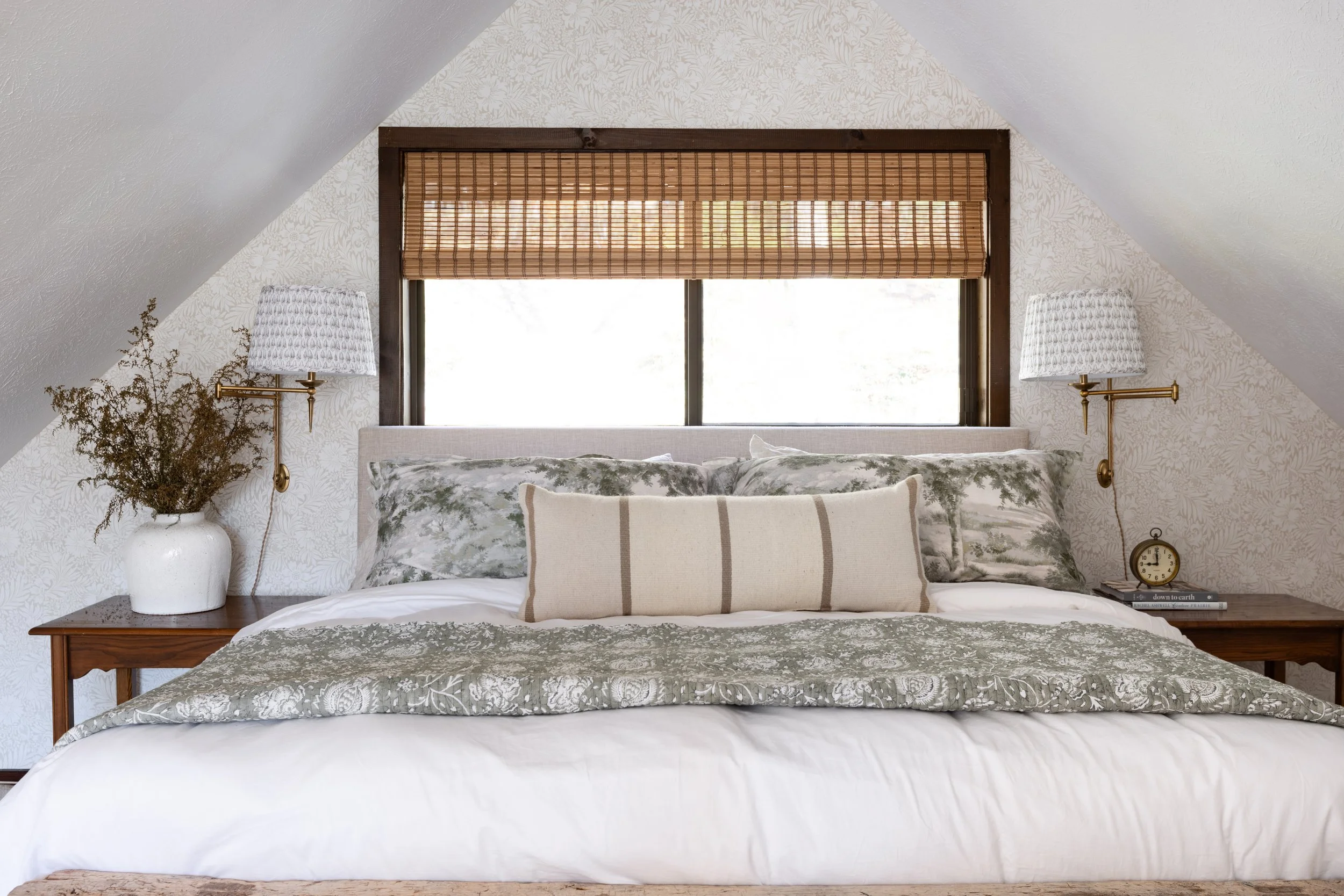 A cozy bedroom with a sloped ceiling, featuring a window with a bamboo shade, a bed with floral and striped pillows, two wall-mounted lamps, a large white vase with dried branches, and a bedside clock and books.