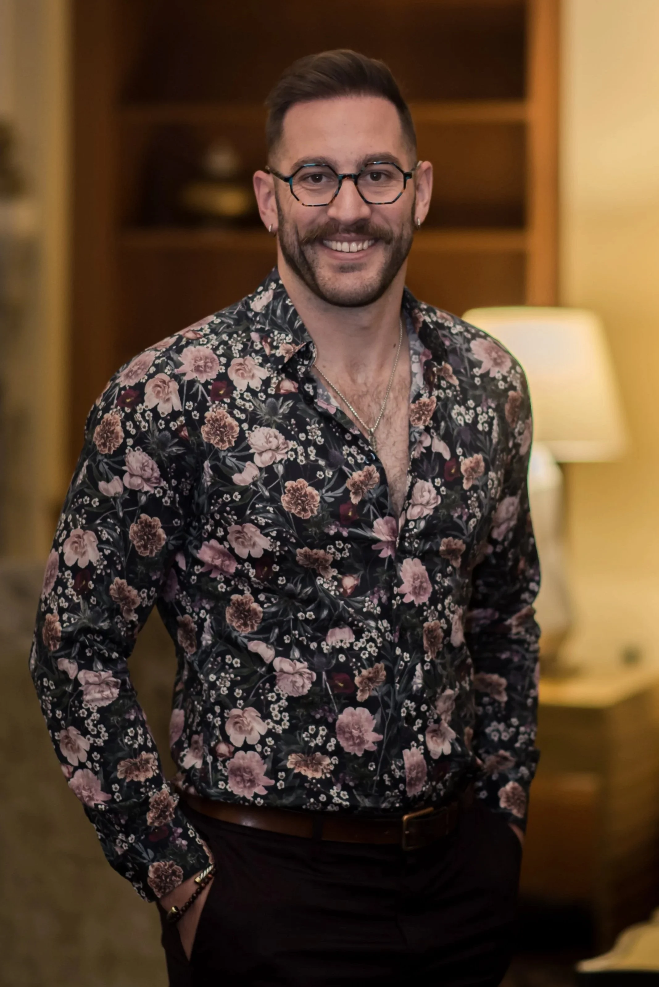A smiling man with glasses and earrings, wearing a floral shirt and black pants, stands in a warmly lit room with shelves and a lamp in the background.