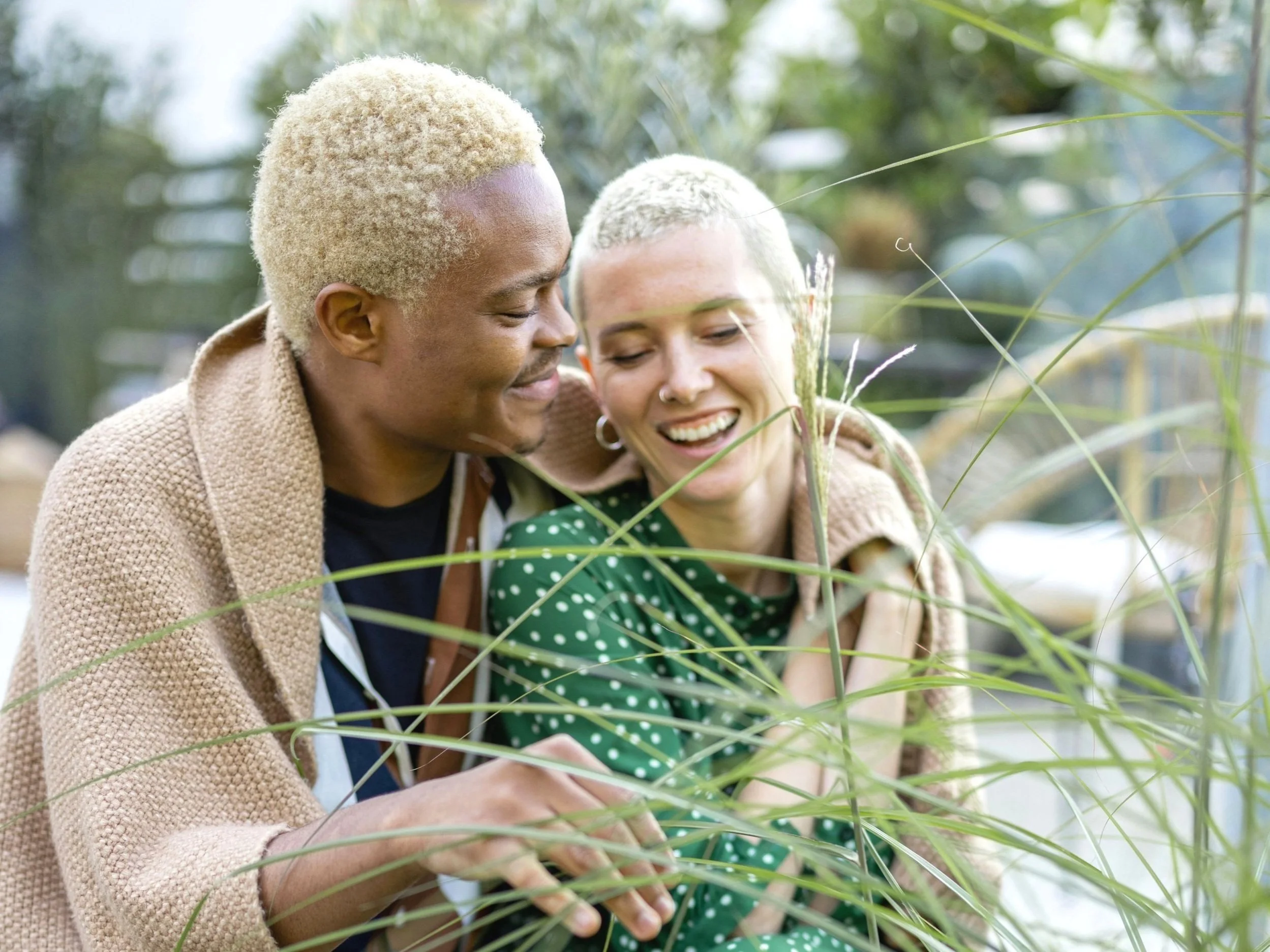 Two people with short blonde hair smiling and enjoying nature, surrounded by green foliage, covered with a brown blanket.