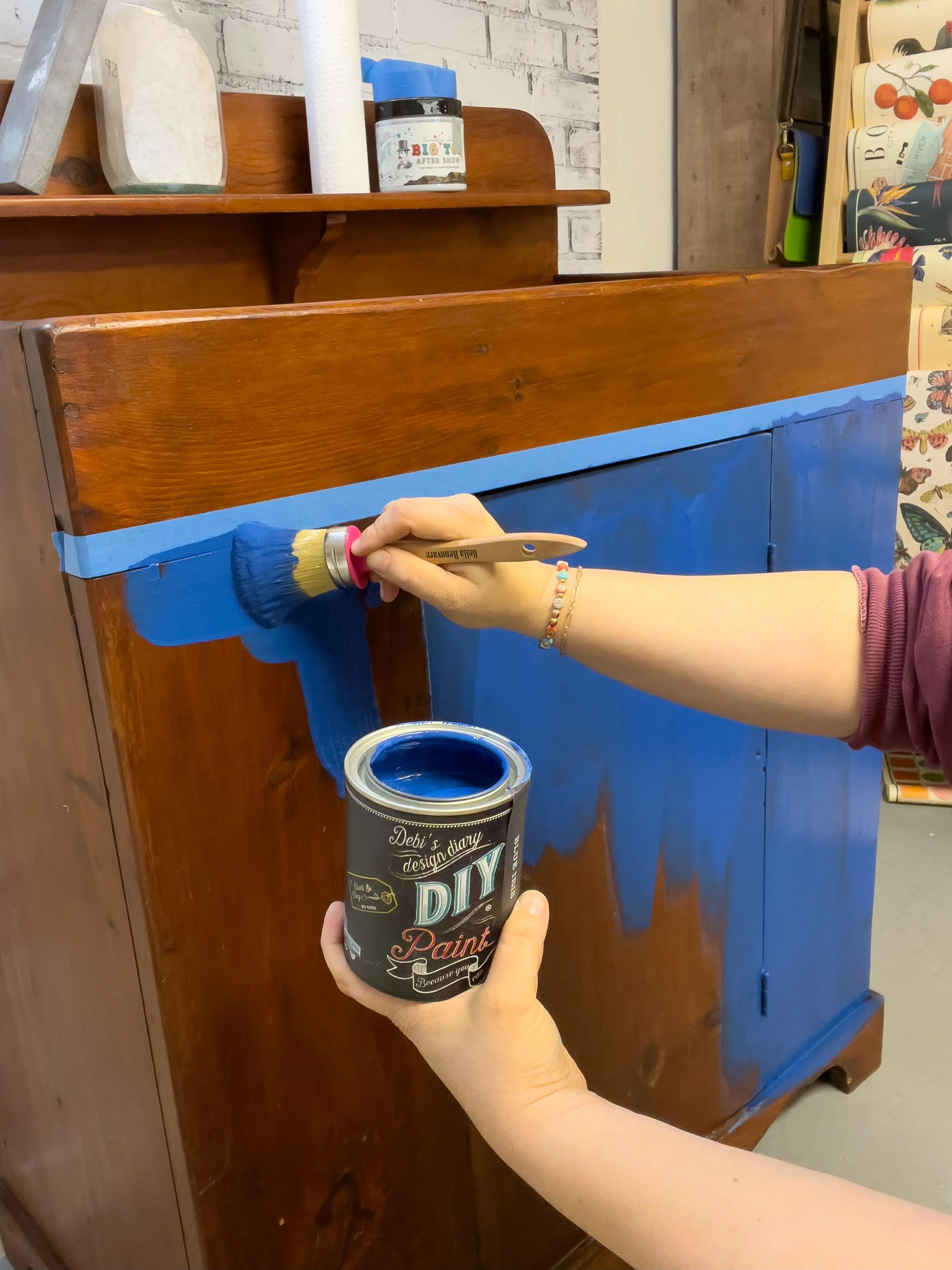 Person painting a wooden cabinet blue using a brush and a can of DIY paint.