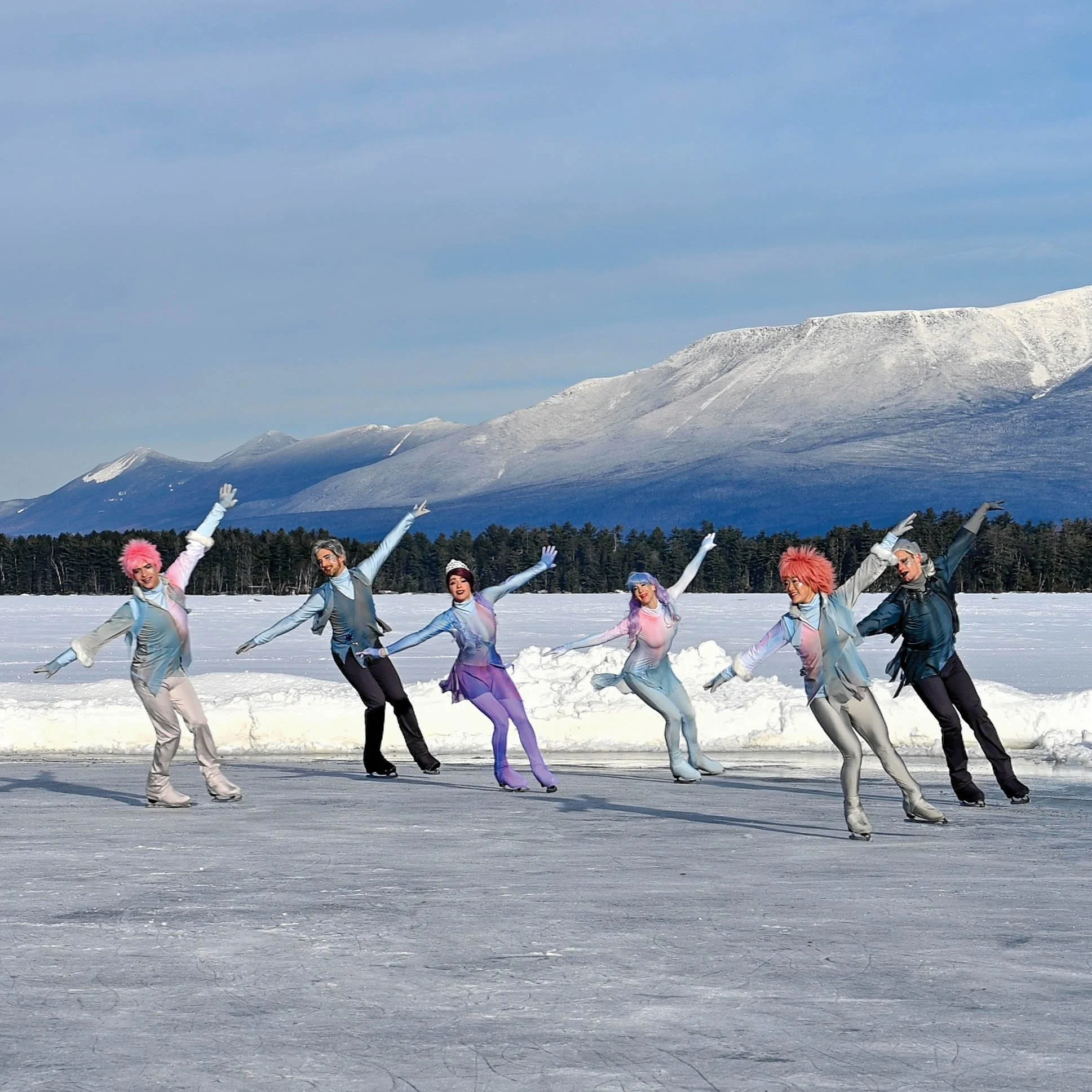 Line of ice skaters on outdoor rink with mountains in the background.